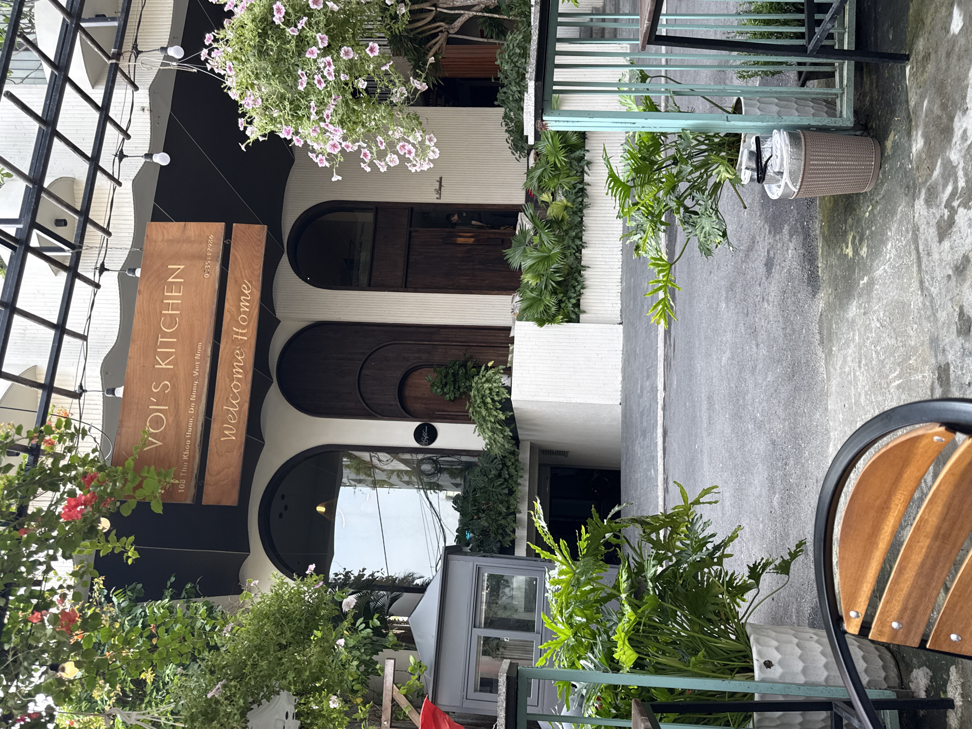 The image captures the exterior of Voi's Kitchen, a restaurant located at 108 Thu Khoa Huan in Da Nang, Vietnam. The restaurant's facade features dark wood doors under rounded archways, and a wooden sign reads "VOI'S KITCHEN" and "Welcome Home." A variety of potted plants and greenery decorate the entrance and surrounding area. A section of outdoor seating, including chairs and metal railing, is visible. The street pavement in front of the building is grey. Above the restaurant entrance, there is a metal-framed canopy, and a string of lights is suspended beneath it. The photograph is taken from an outdoor vantage point, likely from a table or seating area opposite the restaurant.