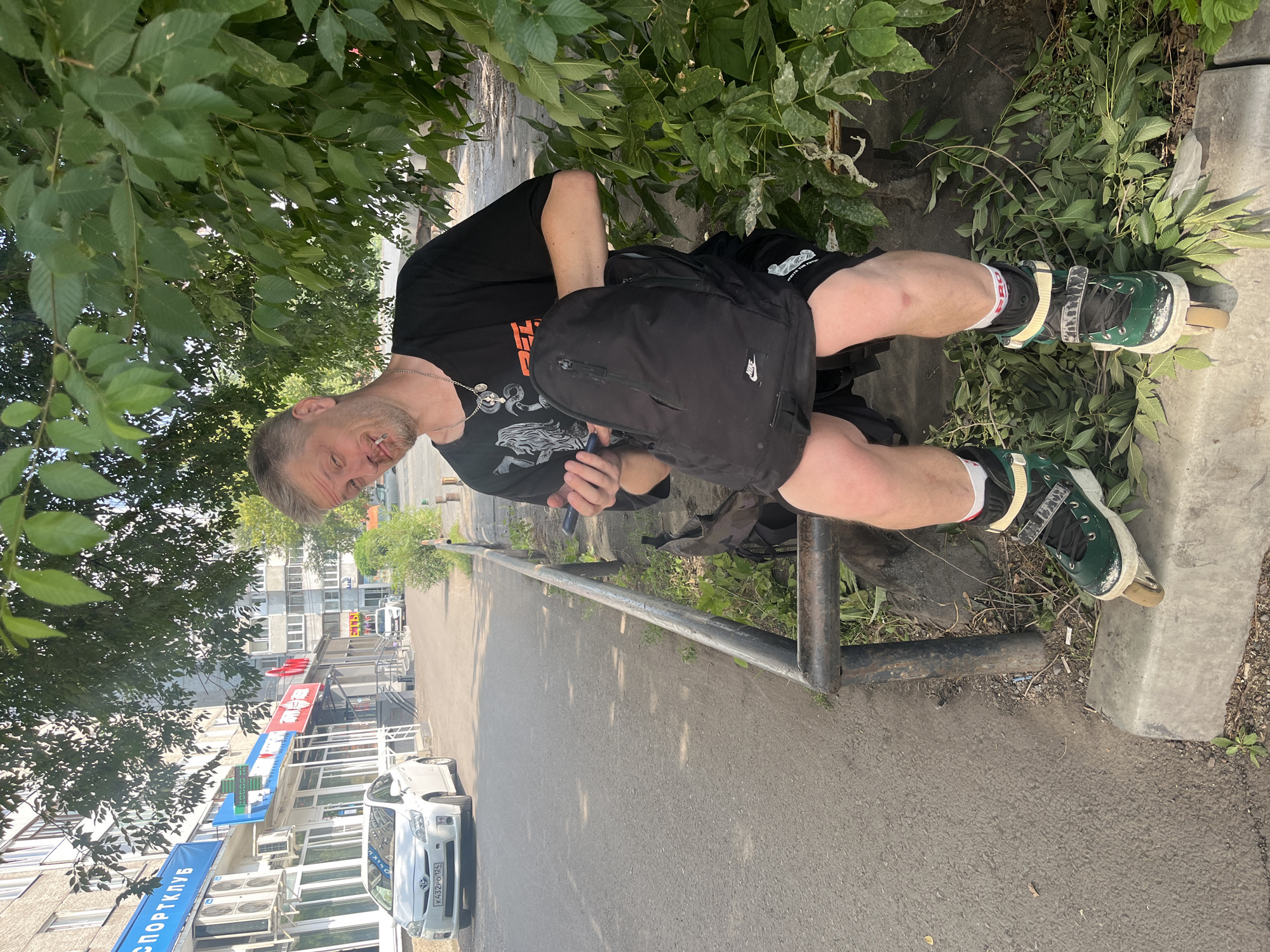 The image shows a man sitting on a concrete curb near a street in Krasnoyarsk, Russia. He is wearing rollerblades, a black t-shirt, and black shorts. A black backpack is on his lap, and he is holding a mobile phone in his right hand. A cigarette is visible in his mouth. The background shows a street with cars and buildings, including a store with a "Спортклуб" (Sport Club) sign. Trees and foliage are present around the man.