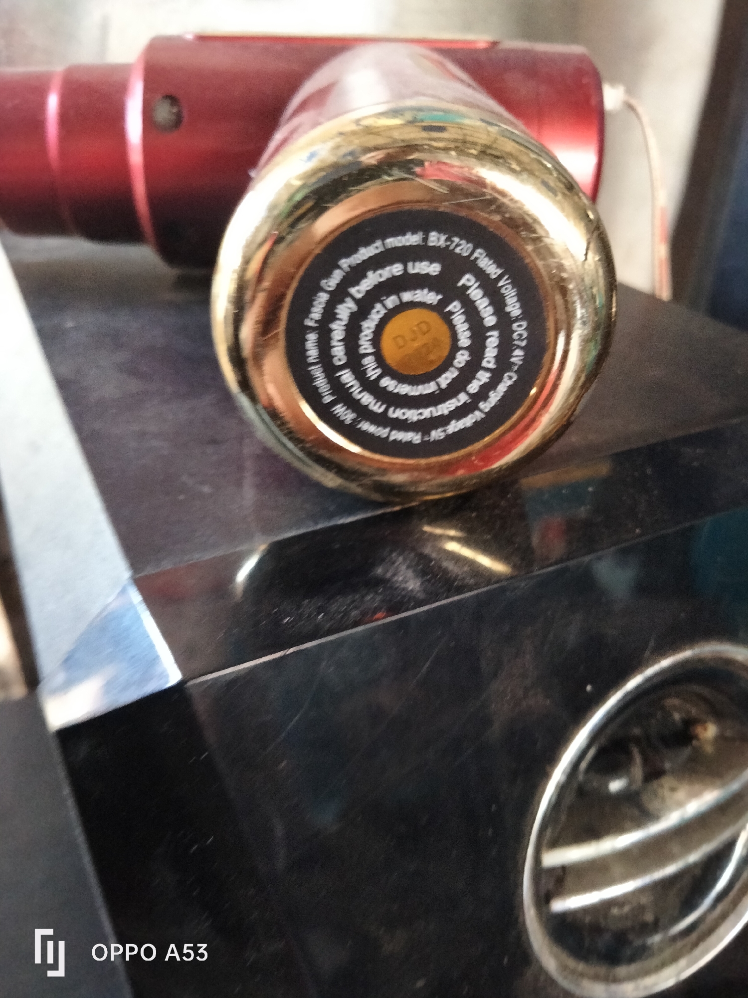 The image displays a close-up, low-angle shot of a red and gold electronic device resting on a dark, textured surface. The device, which appears to be a massage gun, is partially visible in the upper left quadrant, with its red body and a gold circular base filling the foreground. The gold base has text printed around its circumference and a circular yellow label with black text in the center. The text on the gold base is difficult to read due to its small size and the image's resolution, but some phrases like "Product name: BX-720," "Please read the instruction," "do not immerse," "before use," and "DC7.4V" are discernible. The overall environment suggests an indoor setting, likely a home or workshop, given the presence of the electronic device and its placement on a surface that could be furniture. The lighting is ambient, with reflections visible on the shiny surfaces of the device. A watermark in the bottom left corner reads "OPPO A53," indicating the device used to capture the image. The scene is static, with no apparent human presence or activity. The background is blurred, focusing attention on the device. The image does not provide any specific information regarding its location in Denpasar, Indonesia.