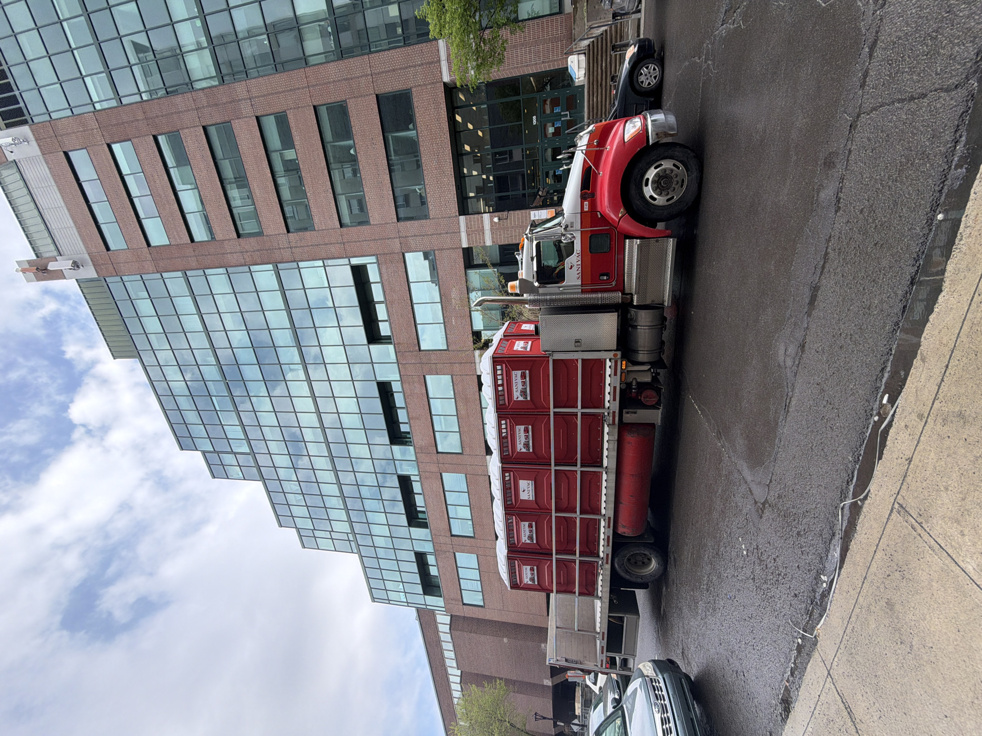 A truck carrying stacked portable toilets is parked on a city street, likely in Montreal, Canada, based on the architecture. The truck is red and white, and the toilets are red with white tops, labeled "Sanivac". Behind the truck is a multi-story building with both glass and brick facade elements. There is a sidewalk on the right side of the image, and the road is wet. Other vehicles are parked nearby. The sky is partly cloudy.