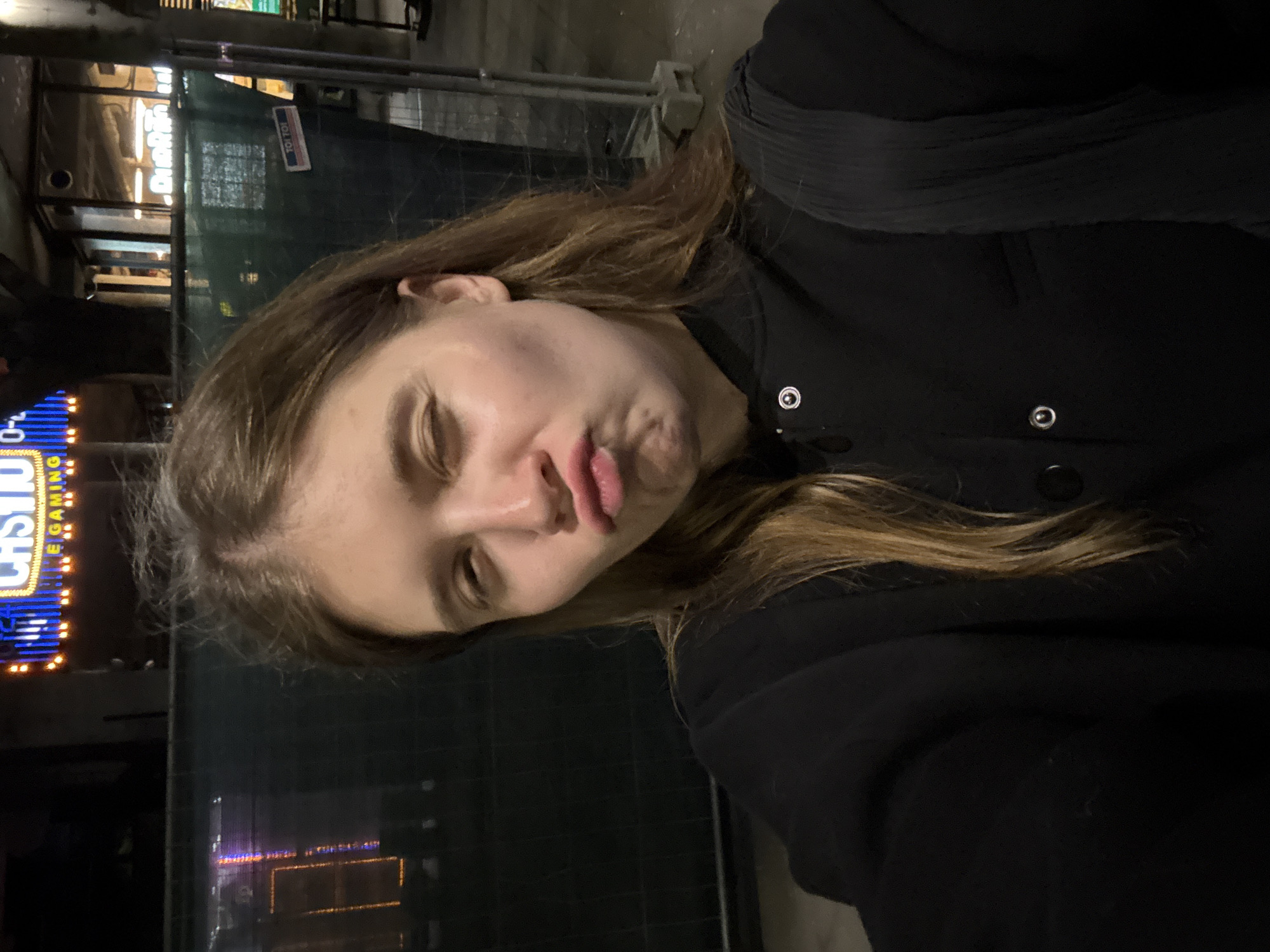 A young woman is captured in a close-up, slightly angled shot at night. She is wearing a dark-colored jacket with button details. Her hair is light brown and appears casually styled. She has closed eyes. In the background, neon signs are visible, suggesting an urban environment. A partially obscured sign that reads "CASINO" is also seen. The location is in the city of Belgrade, Serbia.