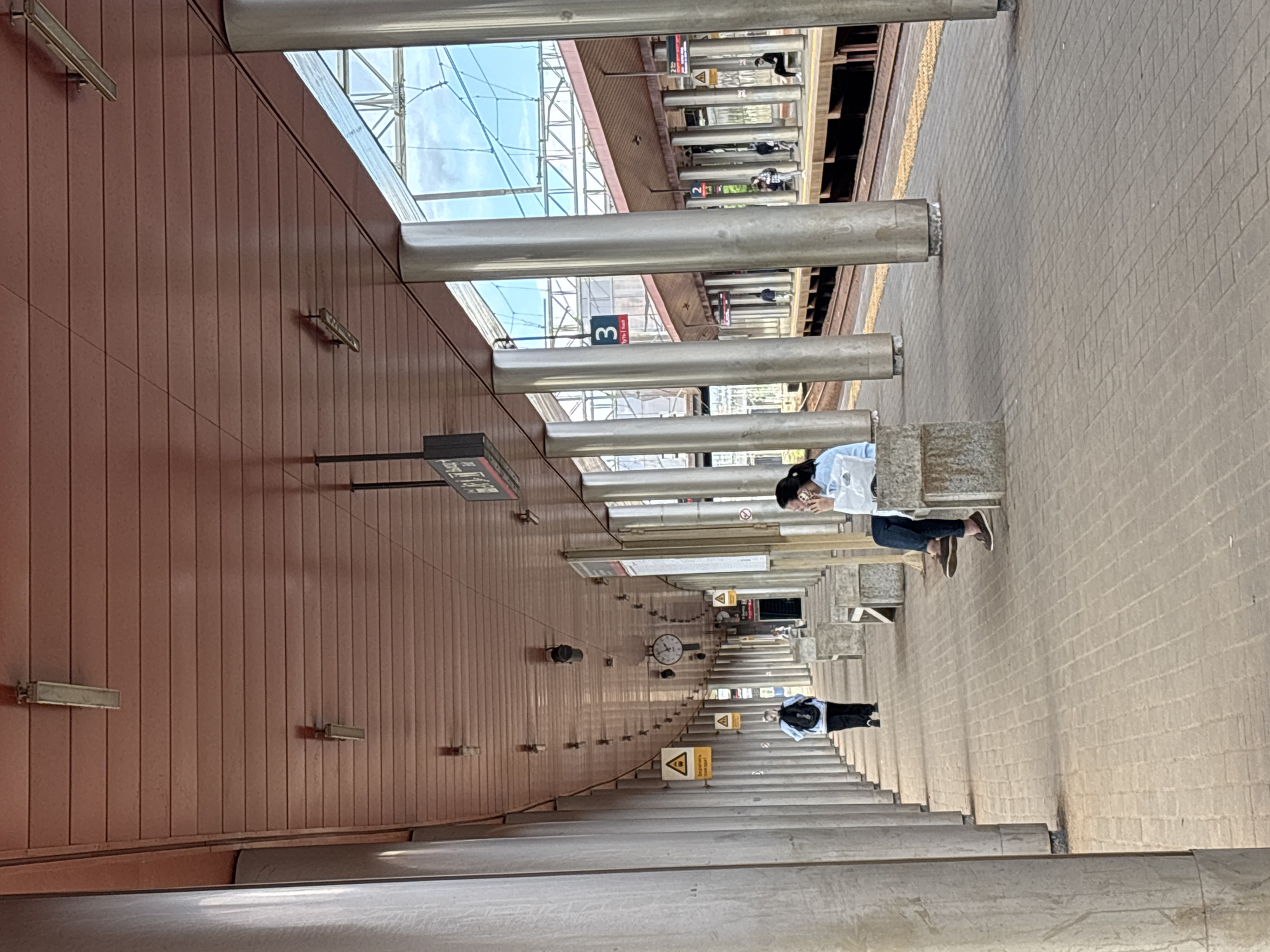 The image depicts the platform of a Moscow Metro station in Russia. The platform is long and straight, with a high ceiling supported by a series of cylindrical columns. A person wearing a white shirt and jeans is seated on a platform bench. Another person, dressed in black, stands further down the platform, appearing to wait. The platform floor is paved with rectangular stones. Warning signs are visible along the platform. The walls are clad in brown panels. A clock is mounted on the wall. The platform is marked with a "3" in a red square, likely indicating the platform number. Overhead are the railway tracks.