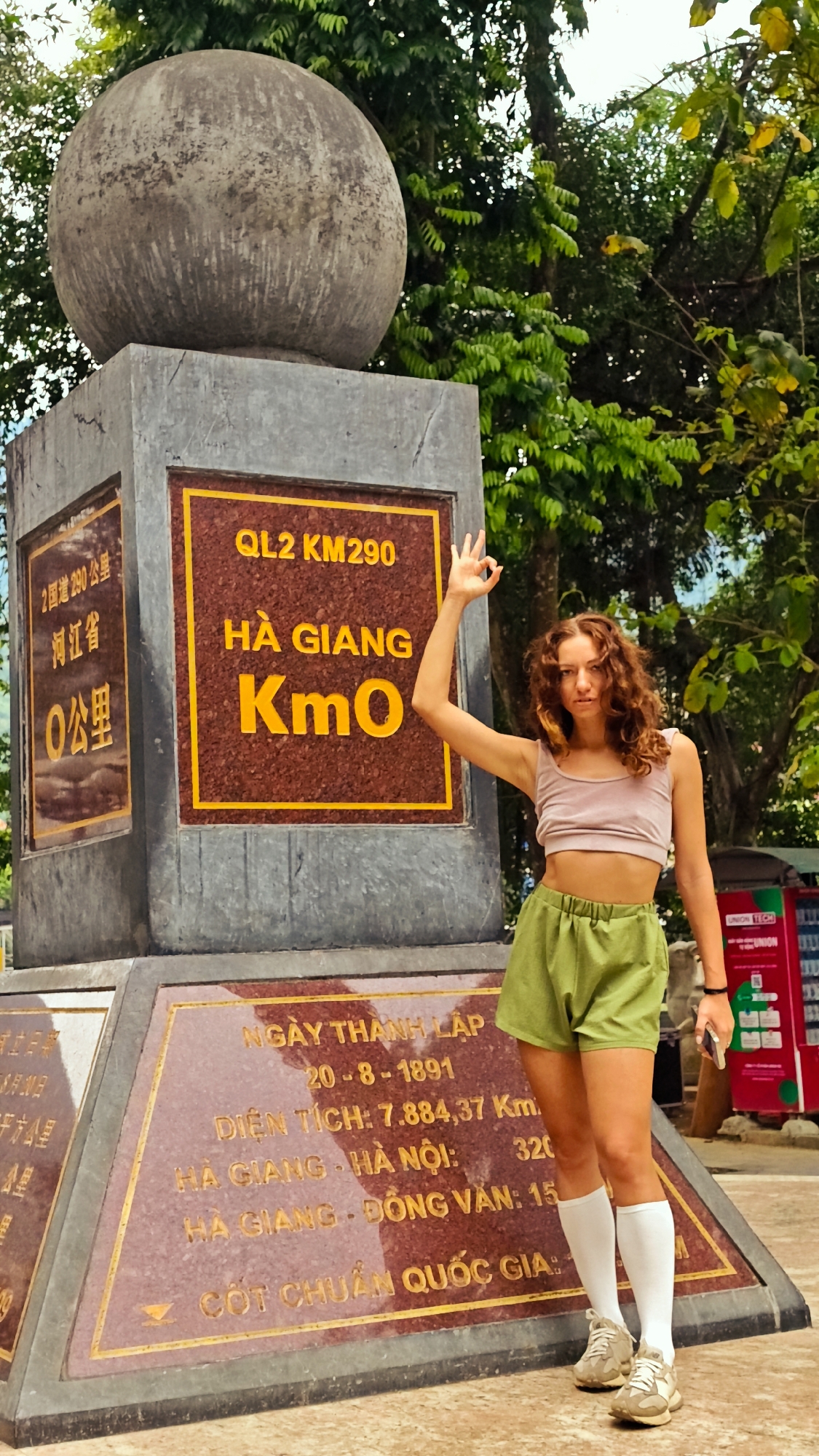 A full, eye-level shot showcases a woman standing next to a monument in Hà Giang 1 Ward, Vietnam. The monument is topped with a large gray sphere. Below the sphere, a gray rectangle displays a sign in yellow and brown that reads "QL2 KM290 HÀ GIANG Km0". Another plaque reads "NGÀY THÀNH LẬP 20-8-1891 DIỆN TÍCH: 7.884,37 Km. HÀ GIANG - HÀ NỘI: 320 HÀ GIANG - ĐỒNG VĂN: 15 CỐT CHUẨN QUỐC GIA". The woman, who has curly brown hair, wears a light pink crop top, green shorts, and white socks with beige sneakers. She raises one hand in a gesture while holding a cell phone in the other. A vending machine is to her right, and green trees form the backdrop.