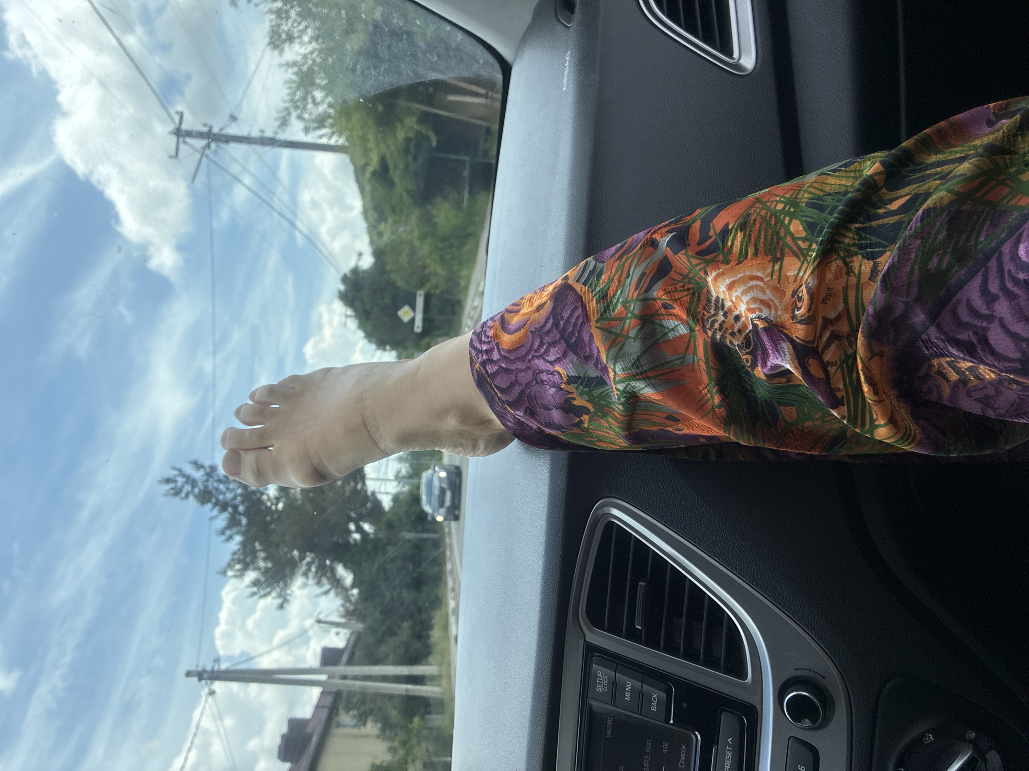 A person's bare foot is resting on the dashboard of a car, with their leg clad in brightly colored, patterned pants. The car's interior is visible, including the radio and air vents. Outside the car, a road, trees, and power lines can be seen under a cloudy sky. In the distance, a dark car is driving on the road. The scene appears to be in a rural or suburban area.