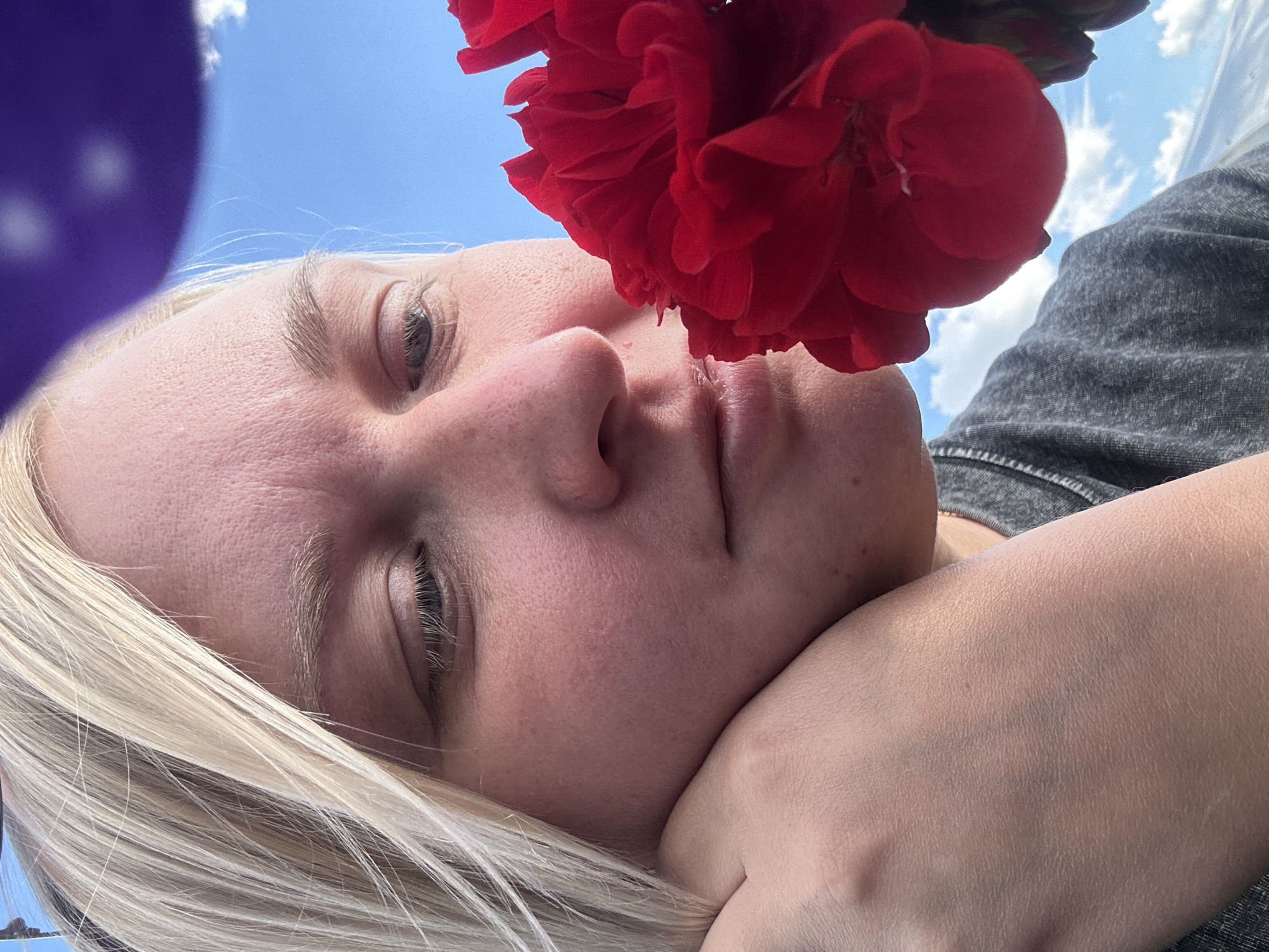 This media file is a close-up, low-angle selfie of a young Caucasian woman lying down outdoors. She has blonde hair that is blowing across her face and is resting her head on her arm. A vibrant red flower is positioned near her face, partially obscuring her features. Her eyes are closed, suggesting a state of relaxation or contemplation. The background shows a bright blue sky with scattered white clouds, indicating it is daytime and likely fair weather. The woman is wearing a dark gray t-shirt. To the left of the frame, a portion of a purple object, possibly a balloon, is visible. The overall mood is serene and peaceful. The image was captured in Chaikovsky, Russia.