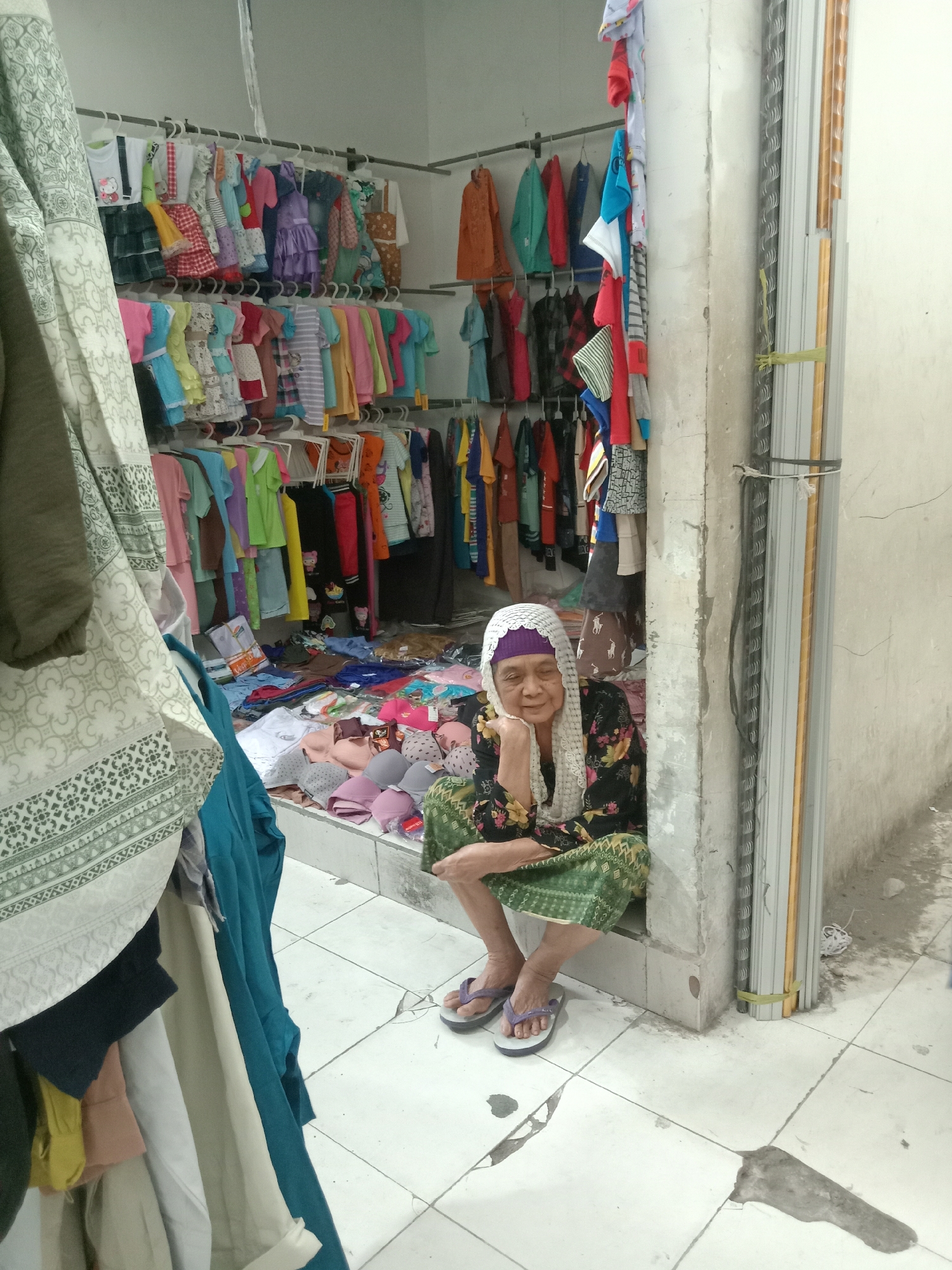 An elderly woman is seated on the floor inside a small shop, possibly a clothing store, in Sugihwaras, Indonesia. She is wearing a patterned long-sleeved shirt, a green patterned sarong, and a white headscarf over a purple head covering. Her left hand is resting on her cheek, and she appears to be looking down with her eyes closed or partially closed. She is wearing flip-flops.
The shop is filled with clothing items, primarily children's clothes, displayed on racks and piled on the floor. Various colorful garments, including dresses, shirts, and skirts, are visible. A stack of bras in different colors is also on the floor in front of the woman. To the right of the woman, a concrete pillar separates the interior of the shop from an outdoor area or alleyway. A stack of metal blinds or shutters is leaning against the pillar. The floor is tiled with white tiles, some of which appear to be stained or cracked. A large patterned cloth is draped on a rack to the left of the frame, partially obscuring the view of the shop's interior. The overall impression is of a modest, cluttered retail space.