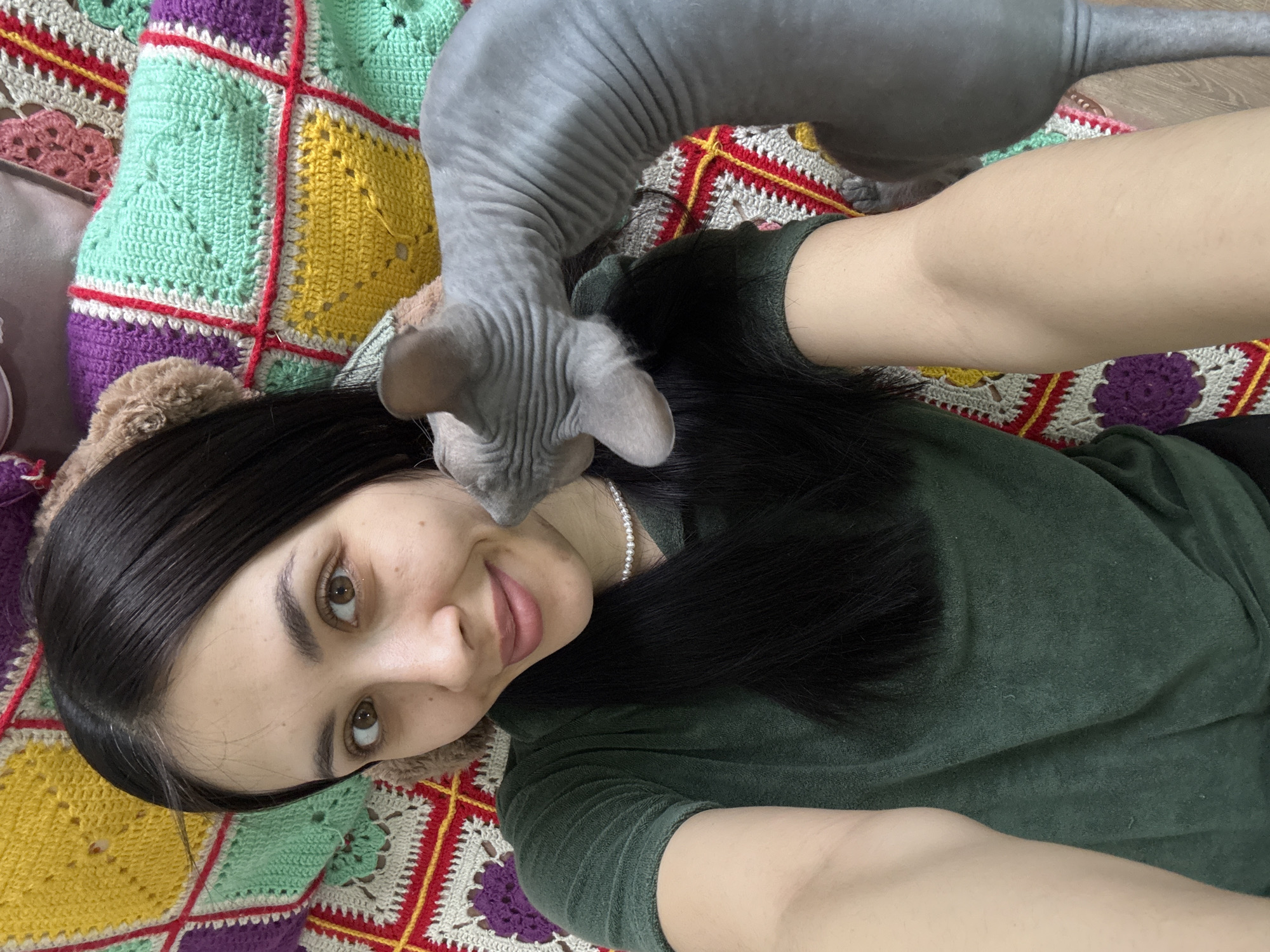 A woman is lying on a multicolored crochet blanket with a sphynx cat on her chest. The woman has dark hair and is wearing a green shirt. She has a pearl necklace. The cat is gray and hairless. The woman is smiling and looking at the camera. The background is blurred. There is no indication of the location being in Votkinsk, Russia.