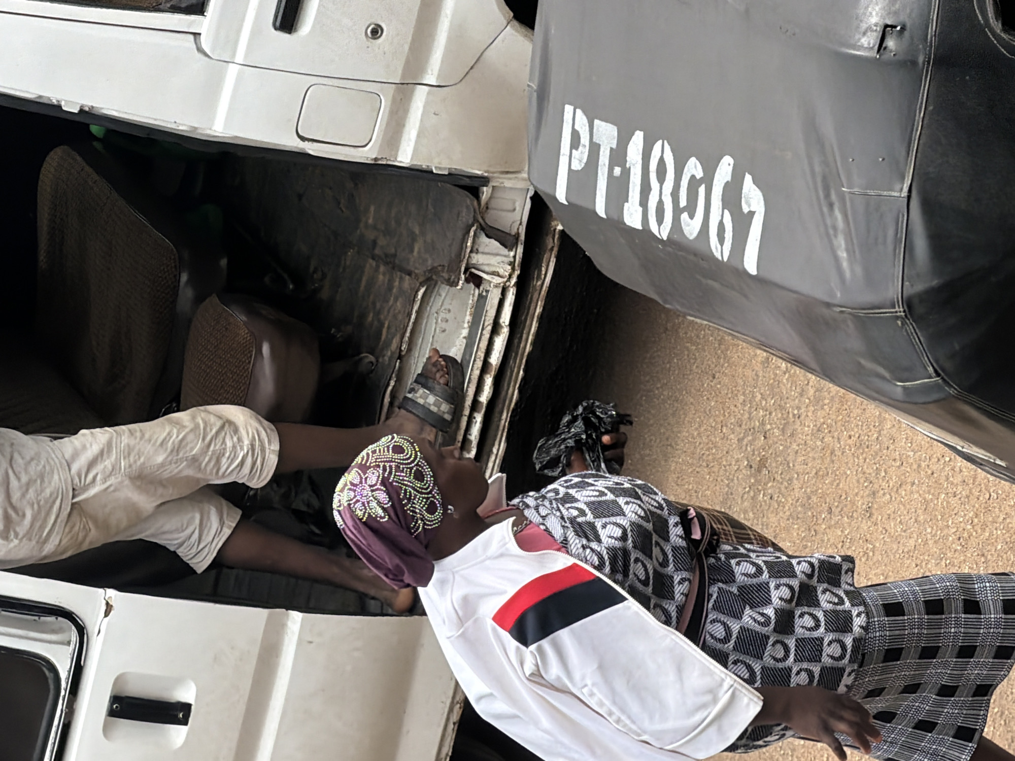 This media file shows an outdoor scene in Bukuru, Nigeria. The primary subjects are two people, seemingly a woman and a child, and a white van. The woman is wearing a purple headscarf with intricate beaded patterns and a white jacket with red and black stripes on the sleeve. She is partially obscured by a patterned black and white blanket or cloth she is holding. A child, wearing white pants, is seated in the open doorway of the van, with their legs visible. The van appears to be old and possibly used for transport, with worn seats visible inside.
The scene suggests a moment of rest or transition, possibly waiting or preparing to travel. The lighting indicates it is daytime, and the ground is a dusty, sandy texture, common in many parts of Nigeria. The van has visible text on its side, "PT-18967", stenciled in white. The overall mood is casual, and there are no strong indicators of emotion or specific events taking place, other than the general context of people and a vehicle in an outdoor, likely urban or semi-urban, environment. The composition is vertical, suggesting the image was taken from a slightly elevated or angled perspective.