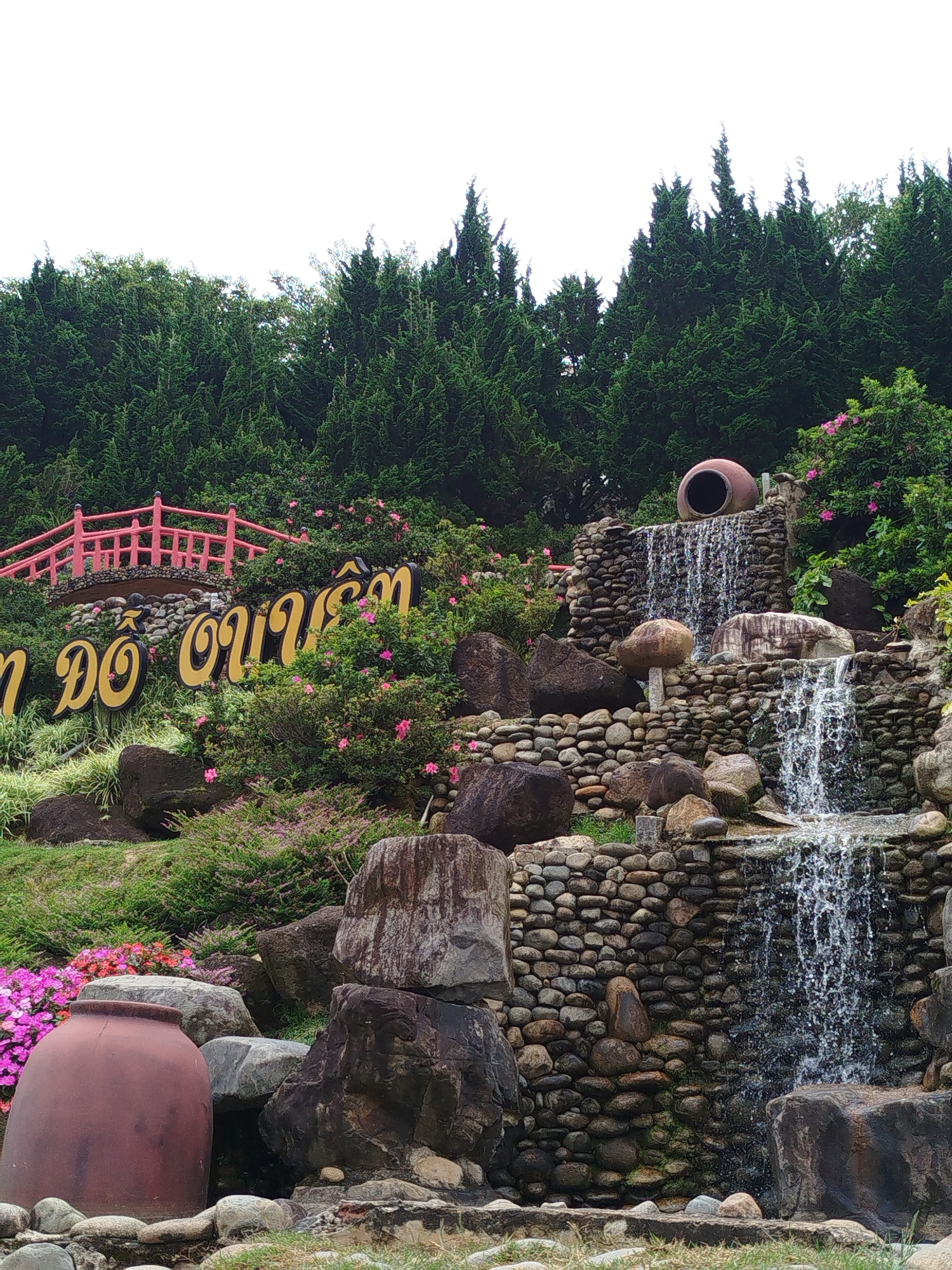 This is an outdoor shot of a scenic waterfall feature, likely part of a tourist attraction, in Lâm Viên - Đà Lạt, Vietnam. The foreground features lush greenery, rocks, and two large clay pots, one a deep reddish-brown and the other a terracotta color. Pink and purple flowers are interspersed among the foliage. A section of a yellow sign with Vietnamese writing is visible, partially obscured by bushes. In the midground, a tiered waterfall cascades down a rock and stone embankment. Above the waterfall, a large, round clay pot sits at the top of the structure, appearing to be the source of the water. A red-railed bridge is visible further up the slope, behind the sign. The background is dominated by a dense wall of dark green evergreen trees, likely cypress or pine, with a bright, overcast sky above. The overall impression is one of a well-maintained, naturalistic garden setting designed for public enjoyment.