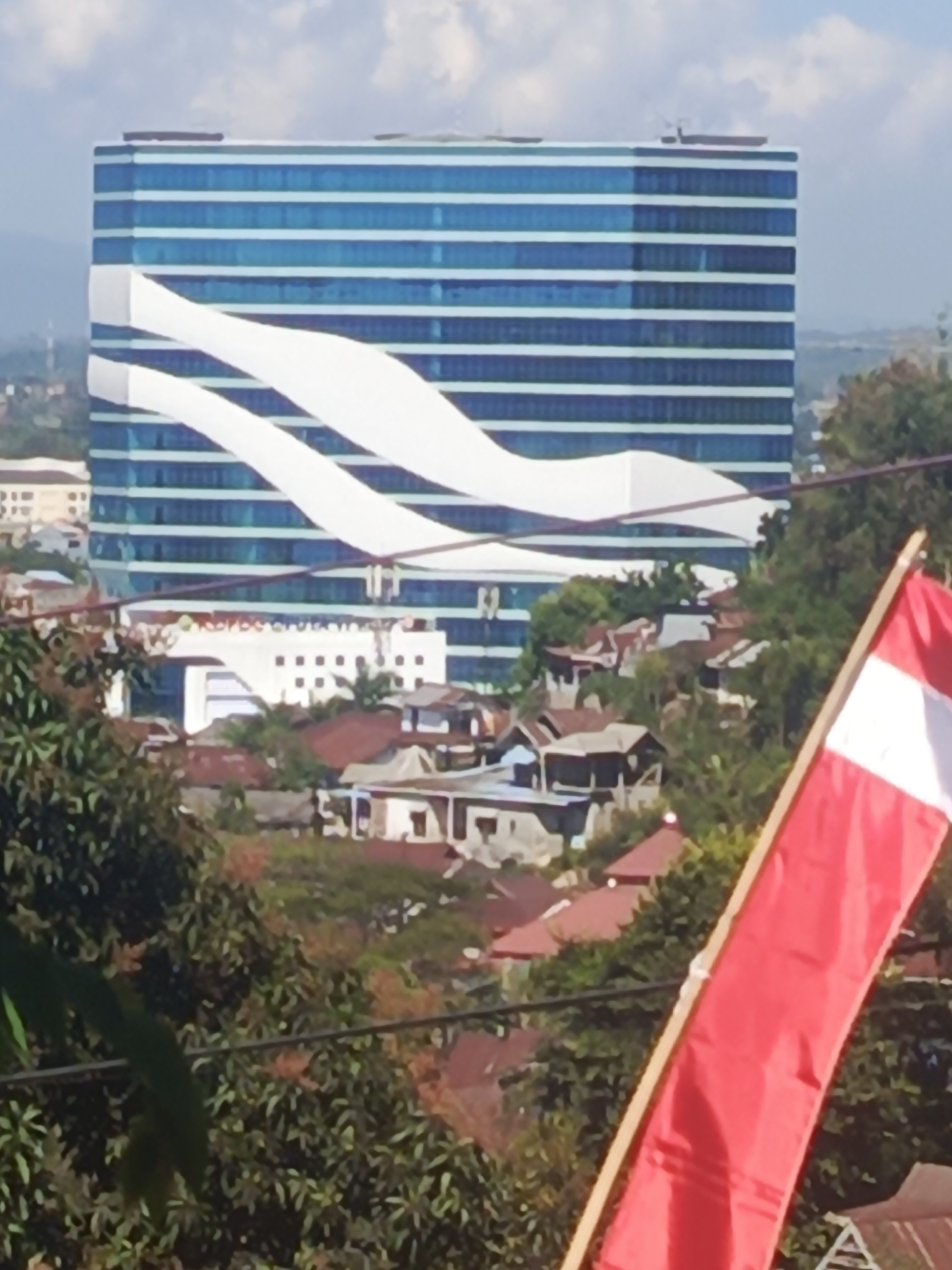 The image shows a cityscape with a modern skyscraper dominating the background. The skyscraper has a distinctive design with horizontal blue bands and flowing white accents that resemble waves. In the foreground, a red and white flag is partially visible, held by a wooden pole. Below the skyscraper, there are several smaller buildings with red roofs, suggesting a residential or commercial area. Lush green trees and foliage are scattered throughout the scene, indicating a natural element integrated with the urban environment. The sky is partly cloudy. Based on the distinctive architecture of the skyscraper, which is known as the Kendari City Hall or Balai Kota Kendari, the image is located in Kendari, Indonesia.