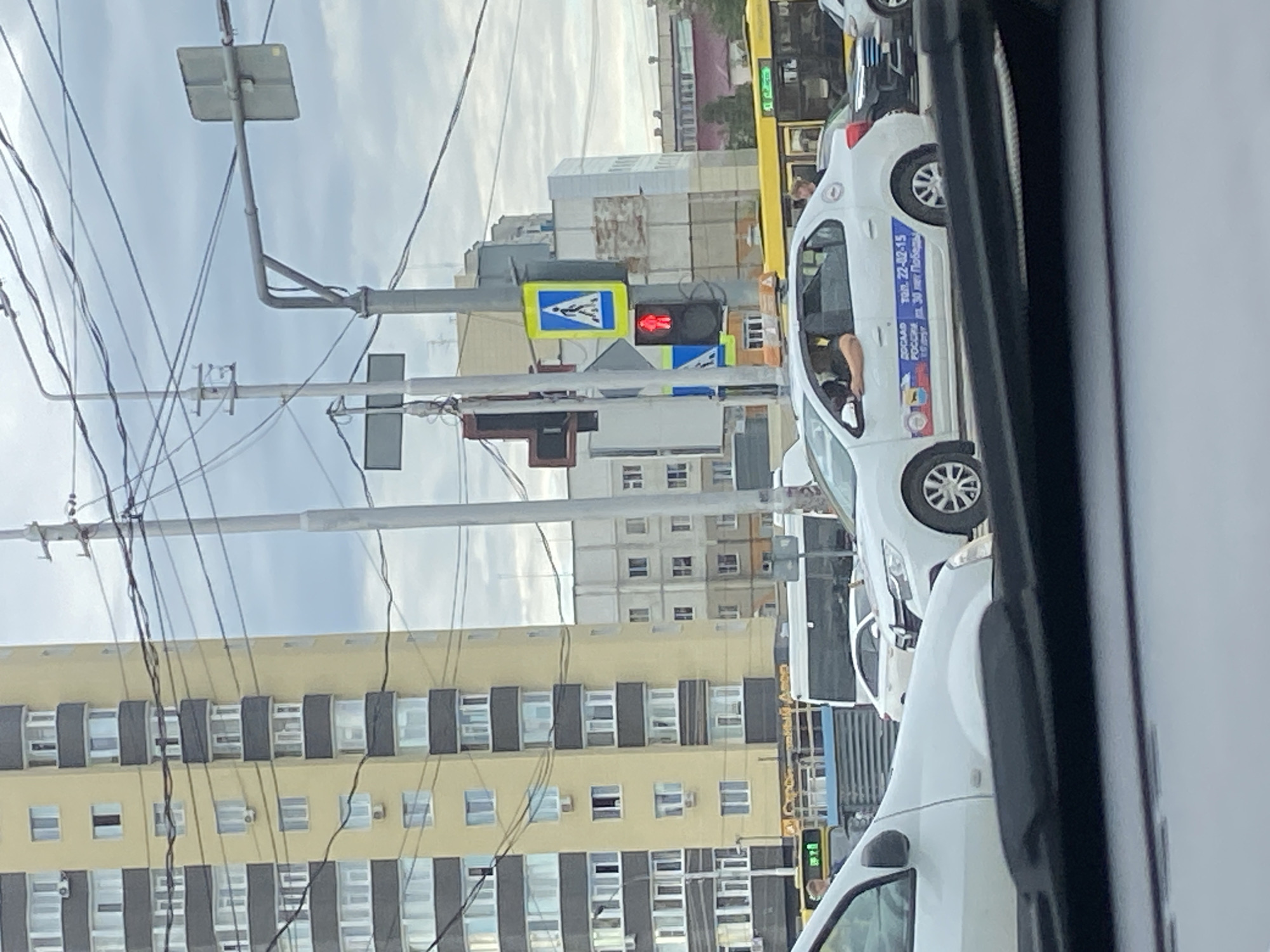 The image depicts a street scene in Surgut, Russia, under an overcast sky. In the foreground, the windshield and dashboard of a white car are visible, obstructing the lower portion of the scene. The main focus of the image is a white taxi, possibly a Renault Logan, with Russian advertisements on its side, driving through a traffic intersection. A person's arm is visible resting on the open driver's side window. Behind the taxi, a yellow bus with advertisements is also present. To the left, a tall, multi-story yellow apartment building dominates the skyline, characterized by its numerous windows and dark balconies. Further in the background, other apartment buildings are visible, some with lighter facades. Traffic lights and electrical wires crisscross the sky, indicating an urban environment. A pedestrian crossing sign with a walking person symbol is illuminated, and a red traffic light is also displayed. The overall impression is a typical day in a Russian city.