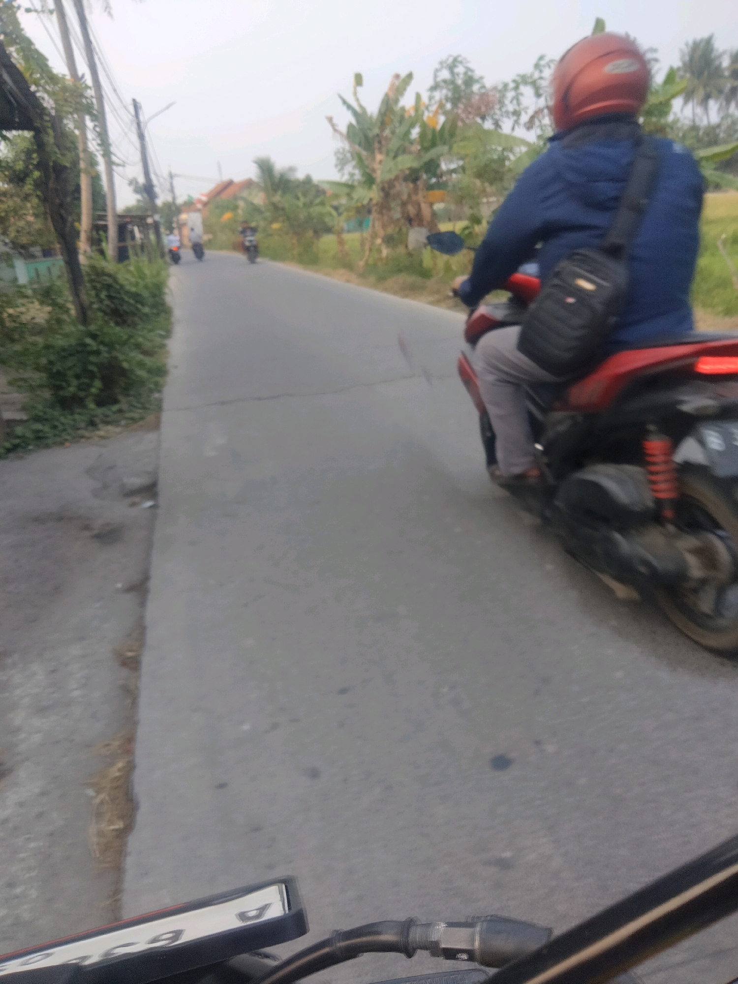 In Suka Sari, Indonesia, on a paved road lined with lush greenery and utility poles, a person wearing a blue jacket and a red helmet is riding a red motorcycle. The rider has a black bag slung over their shoulder, and the motorcycle's license plate is partially visible. The road appears to be a typical rural or suburban street, with trees and other vegetation on either side. The sky is overcast.