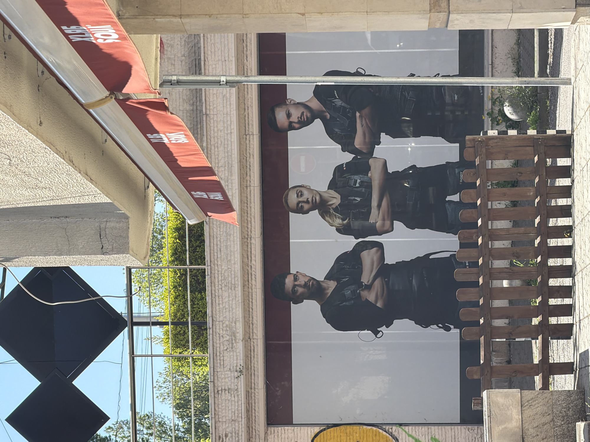 The image depicts a building facade in Sofia, Bulgaria. A red awning with white lettering "XL95 SOUX" is visible at the top left. A large advertisement featuring three people wearing dark clothing and tactical vests is displayed on the building. A wooden fence and a small planter are situated in front of the advertisement. A decorative geometric design is attached to the building's facade to the left of the advertisement.