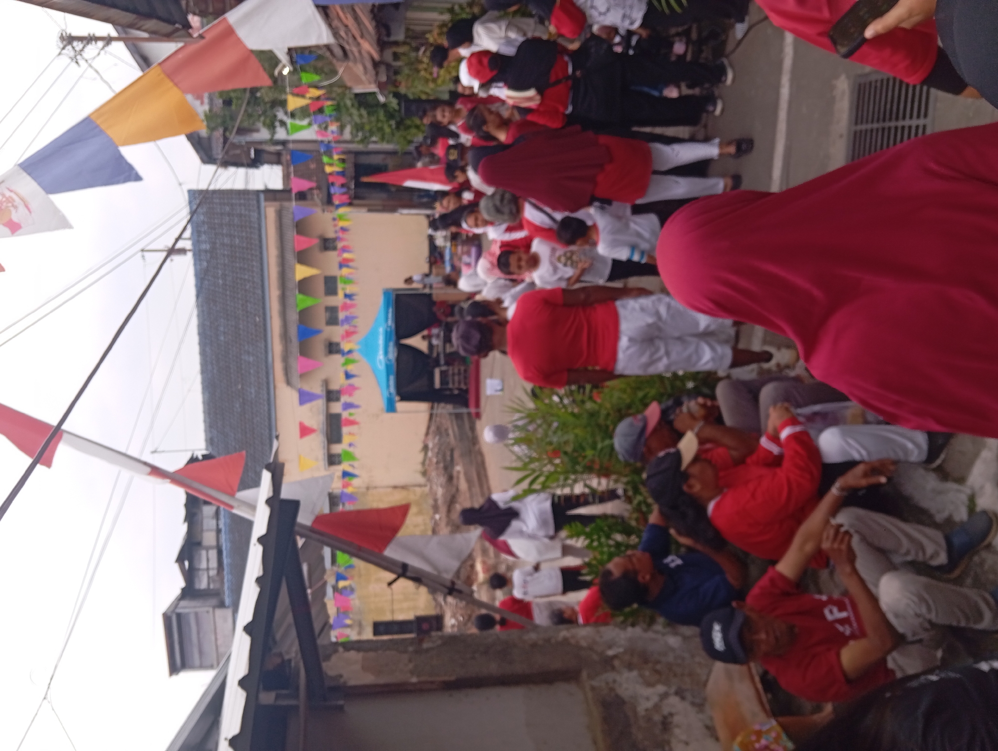 The image depicts an outdoor gathering in what appears to be a residential street in Yogyakarta, Indonesia, possibly during a festive or commemorative event. Numerous people are present, many wearing red or white clothing, which are the colors of the Indonesian flag, suggesting a national celebration.
In the foreground, several individuals are seated, engaged in conversation or observing the surroundings. One man in a red shirt with a "P" logo and a black baseball cap is prominent. To his right, a woman in a red long-sleeved shirt and white pants is visible. Further back, a crowd of people stands and moves around, some holding Indonesian flags.
In the background, a street is lined with buildings, some appearing to be houses. Bunting flags in various colors, including red and white, are strung across the street, adding to the celebratory atmosphere. A blue tent or stage is set up in the middle ground, with individuals standing around it. The sky is overcast, indicating a cloudy day. The overall scene conveys a sense of community participation in a local event.
