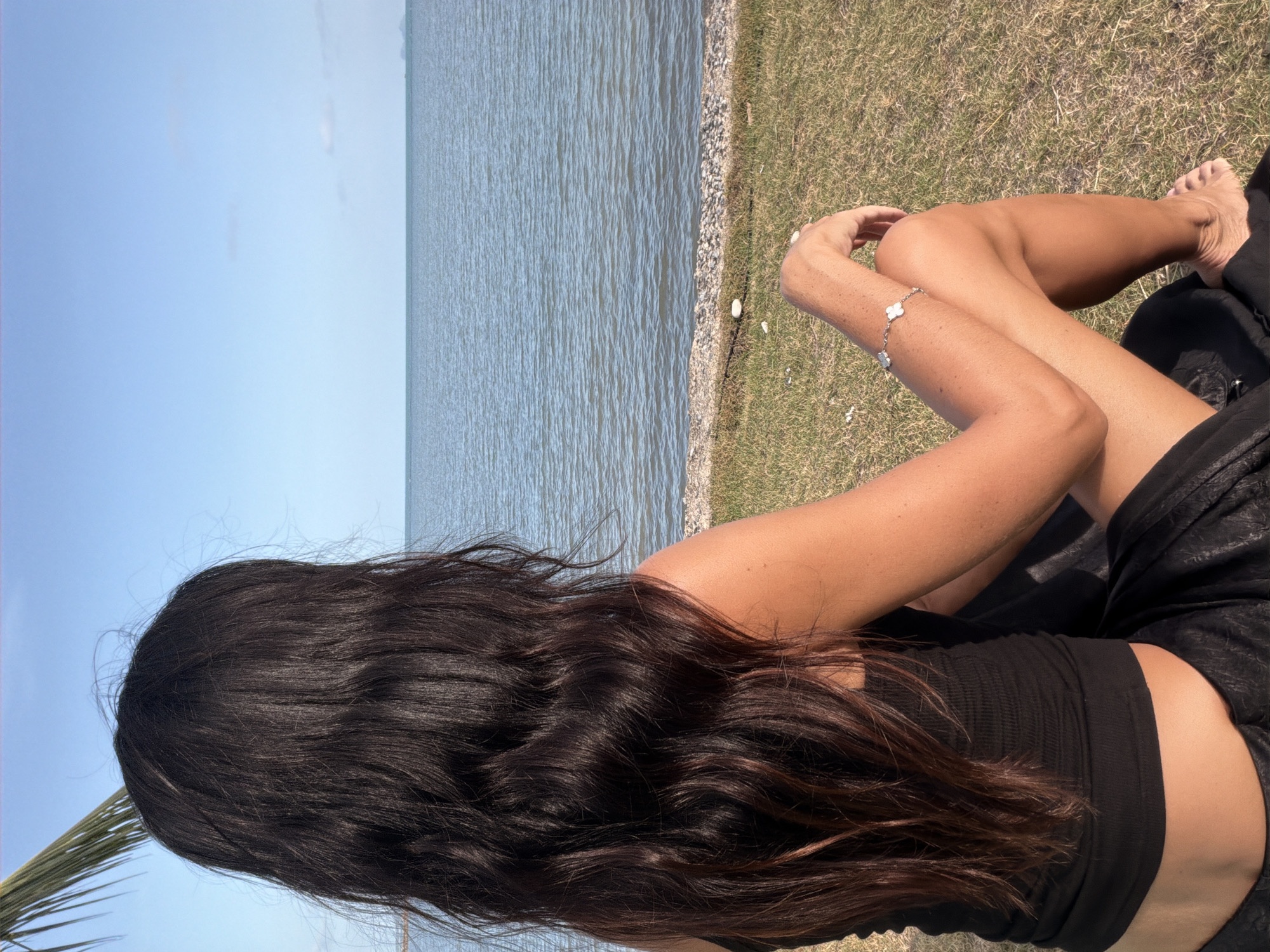 A woman with long, dark, wavy hair sits facing away from the camera, looking out at the ocean. She is wearing a black tank top and a black skirt. Her right arm is resting on her knee, and she is wearing a bracelet with four leaf clover charms. The woman is sitting on a grassy area next to the sea. The sky is a clear blue, and the water is a calm blue-green. The beach is lined with small rocks and pebbles. A palm frond is visible in the upper left corner of the image. The location is identified as Ko Samui District, Thailand, based on the visual cues of a tropical beach and seascape.