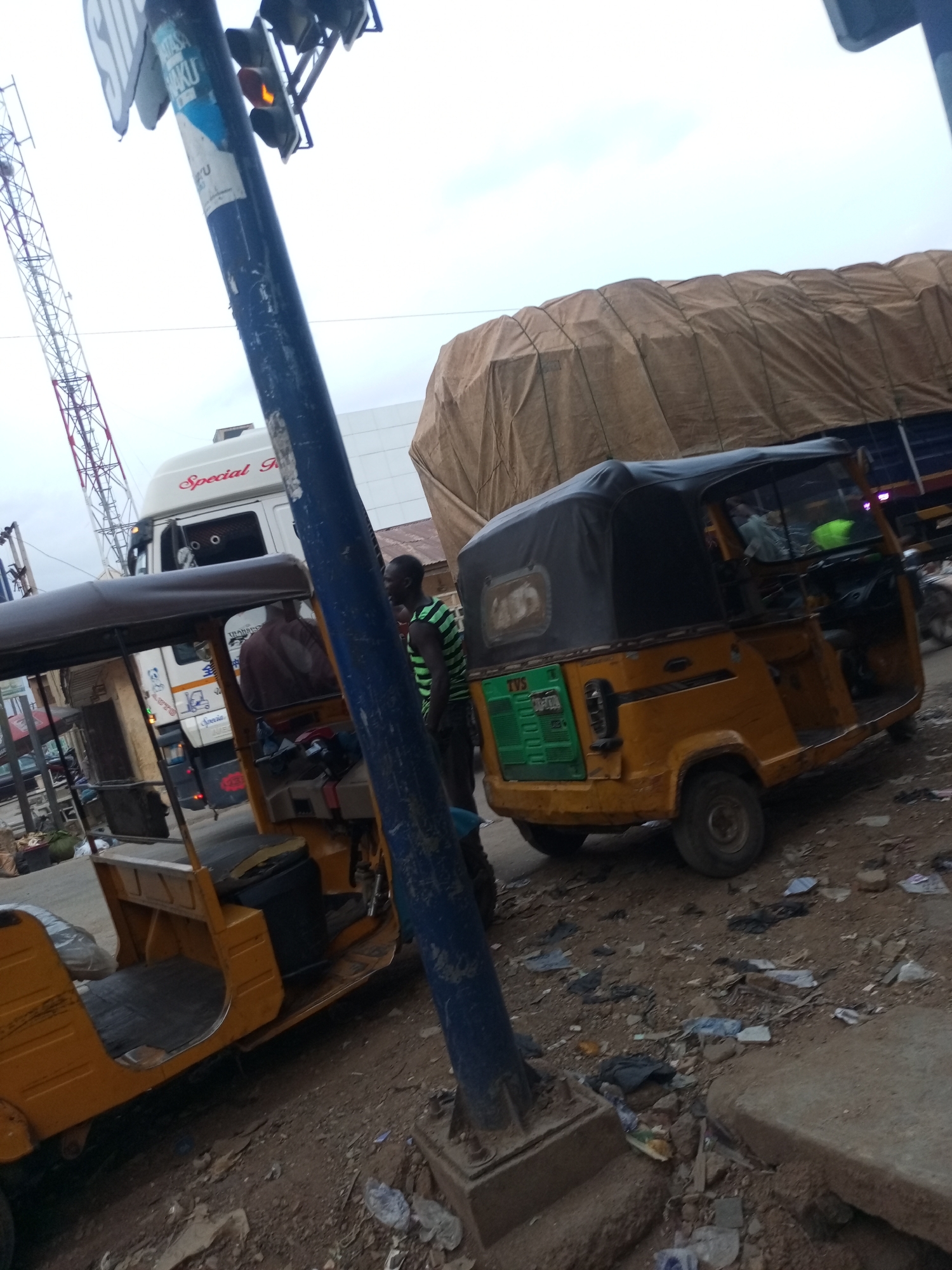 The image shows a street scene in Maiburiji, Nigeria. Two yellow auto-rickshaws (Tuk-tuks) are parked on a dirt and debris-strewn surface. One is partially obscured by a blue utility pole with a traffic light showing yellow. A man wearing a green striped shirt stands near the Tuk-tuk. Behind them is a large truck covered with a brown tarp and a white truck labeled "Special R." A cell tower is visible in the background. The sky is overcast.