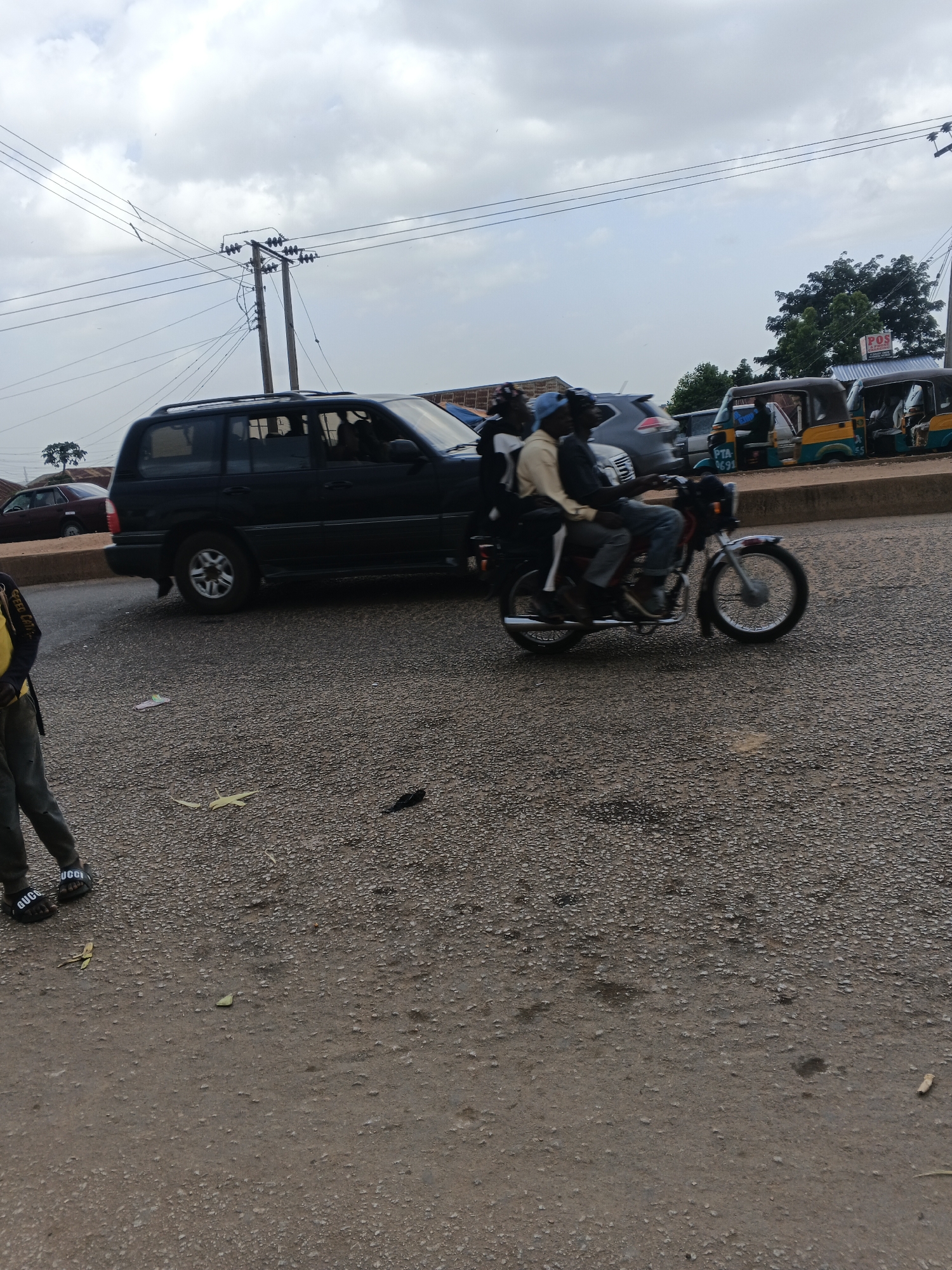 The image shows a street scene in Bukuru, Nigeria. A black SUV is parked on the right side of the road. A motorcycle with two occupants is riding on the road. In the background, there are several auto-rickshaws parked, and a building with a "POS" sign. The ground is covered in gravel and dirt. The sky is cloudy.