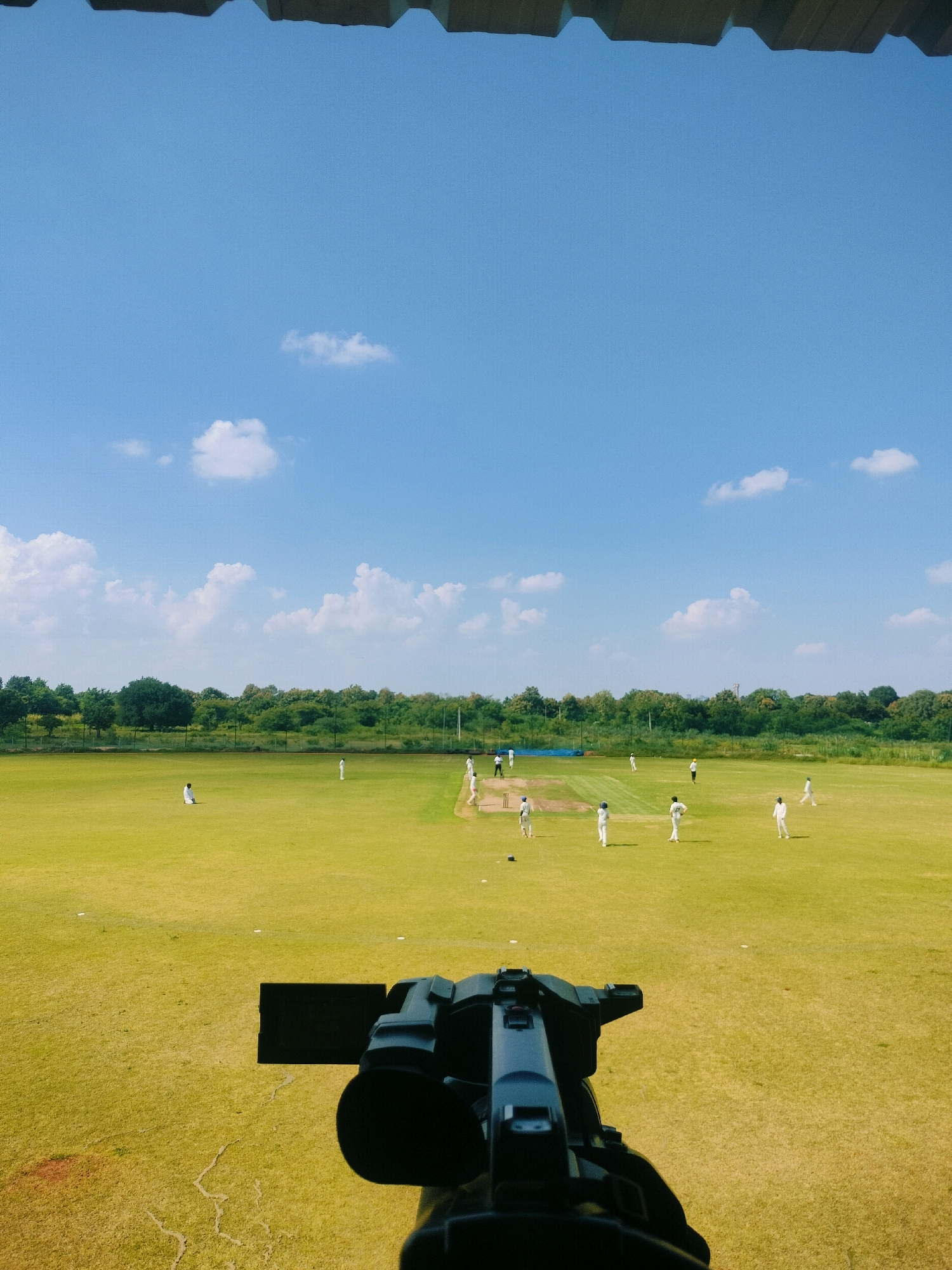 This image depicts a bright, sunny day at a cricket ground in Chilkur, India. The foreground is dominated by a black video camera, positioned at a low angle. Beyond the camera, a vibrant green cricket field stretches out, marked with white lines. Several figures, dressed in white cricket attire, are scattered across the field, engaged in a game of cricket. Some appear to be fielding, while others are positioned as batsmen or bowlers. In the background, a dense line of green trees forms a natural boundary, with a clear blue sky dotted with white clouds overhead. The overall scene suggests a sporting event in progress, captured from a filming perspective.
