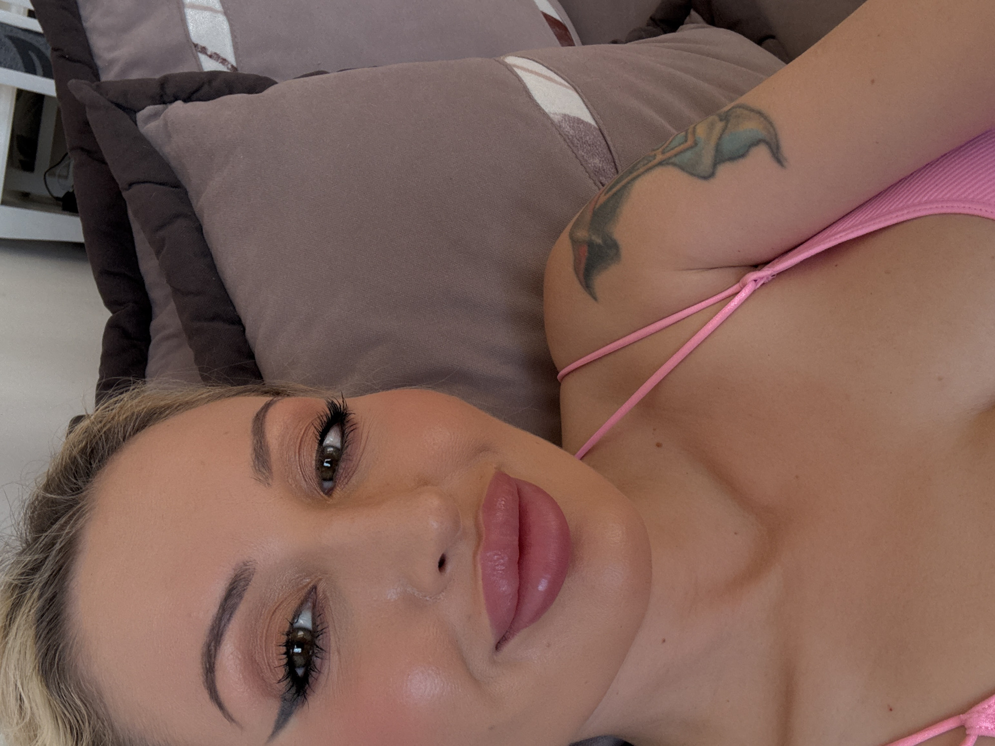 A close-up, top-down selfie shows a woman with blonde hair pulled back, wearing a pink top. She has heavy makeup, including winged eyeliner and pink lipstick. She is lying down, with pillows visible behind her. A tattoo of a bird is visible on her left arm. The lighting is bright and appears to be natural.