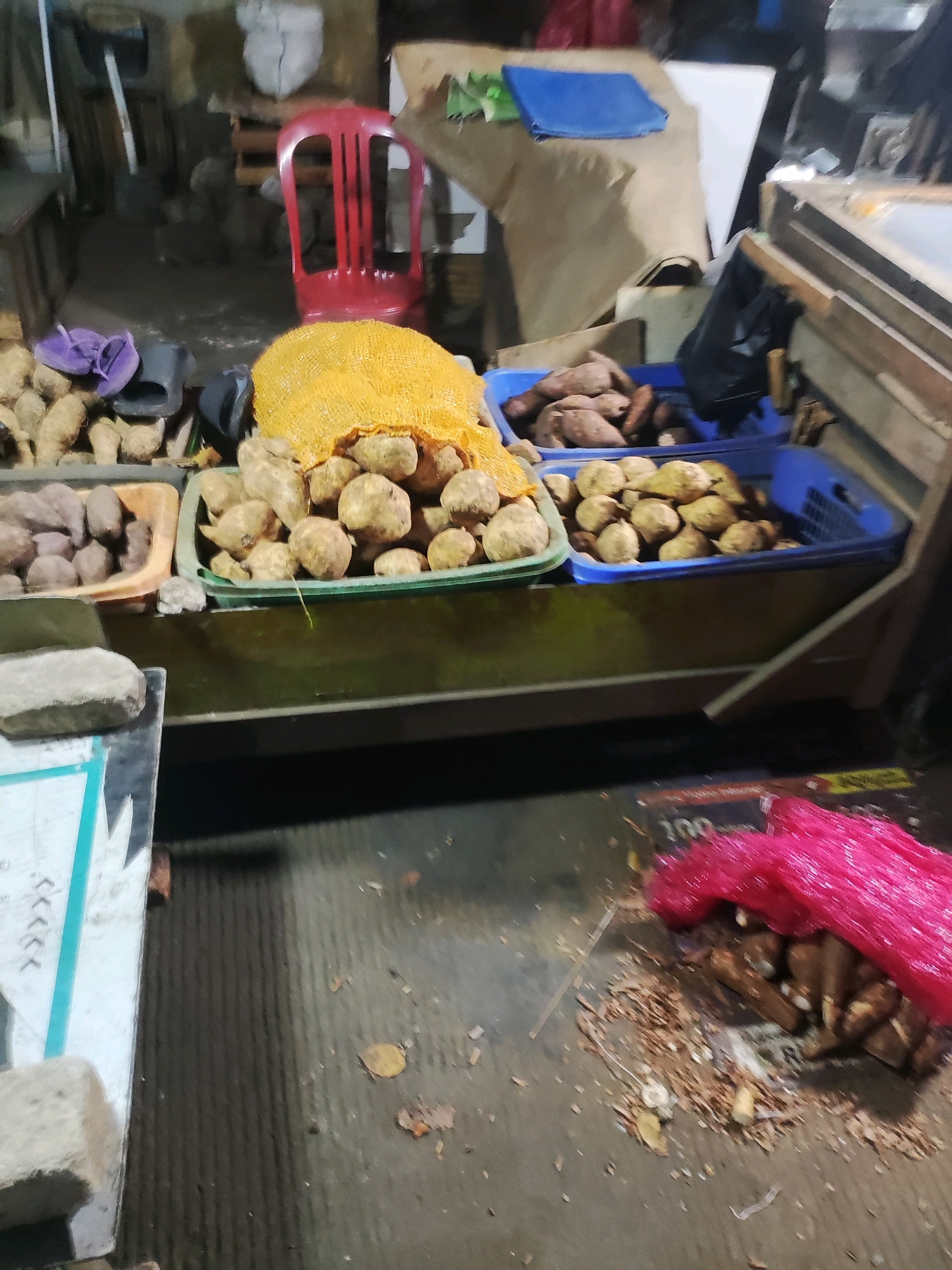 The image depicts a street-level market stall in East Jakarta, Indonesia. The stall features an assortment of root vegetables, likely sweet potatoes, displayed in plastic crates and sacks. In the foreground, a grey, textured surface with a white and teal pattern is visible, possibly a table or stall counter. To the right, a pile of bright pink netting covers more root vegetables scattered on the ground, along with debris such as wood shavings and leaves. In the background, a red plastic chair, a bag of produce, and other market elements are partially visible. The overall environment appears to be an outdoor or semi-covered market setting.