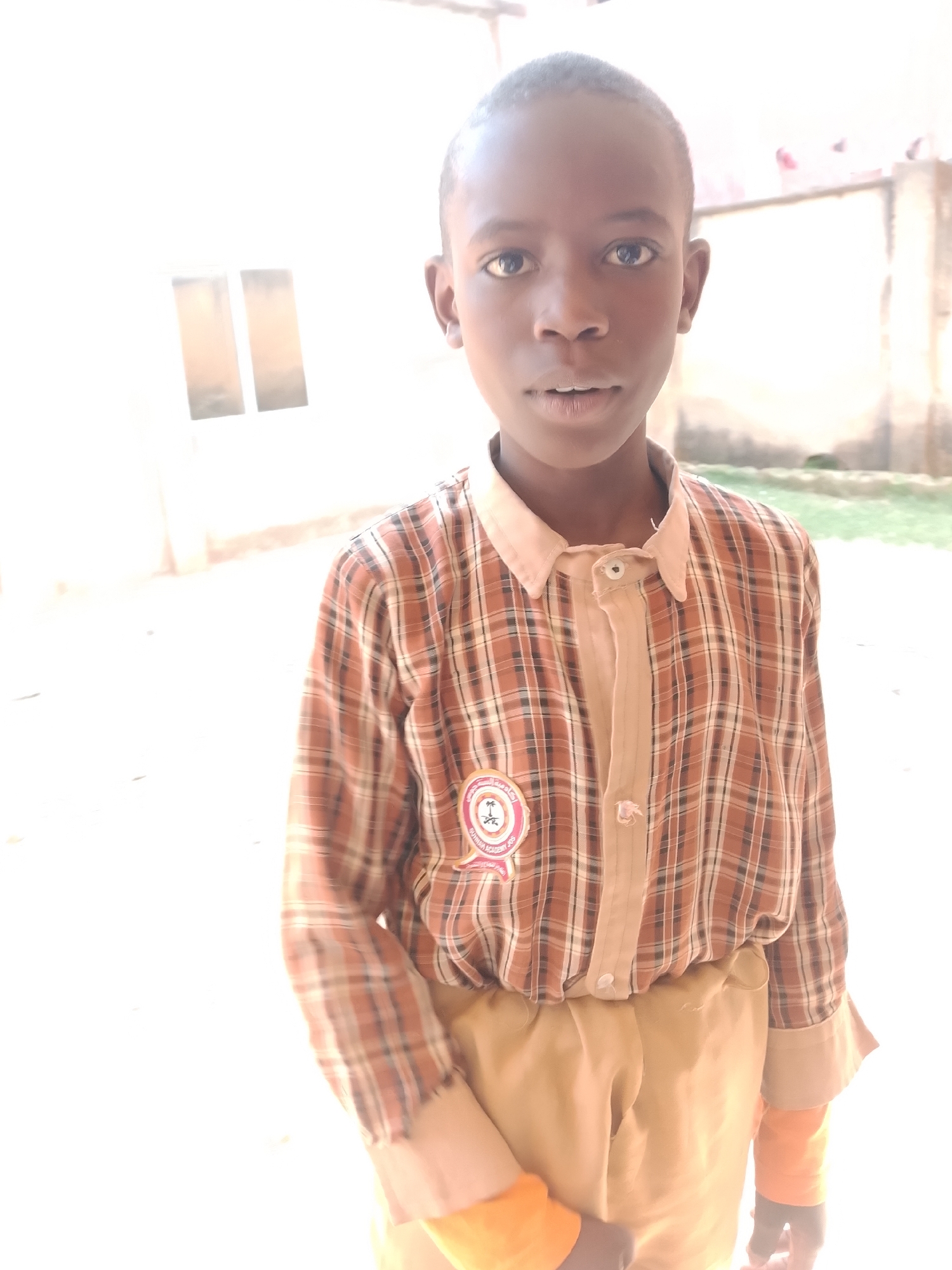 A young Black boy stands in front of a building, looking directly at the camera. He is wearing a plaid shirt with an inner beige collar and a patch on the left side of his chest. His pants are a light brown color. The background shows a white wall with two rectangular windows and some greenery. The lighting is bright, suggesting it is daytime. The scene appears to be outdoors in a courtyard or open area. The location is unknown.