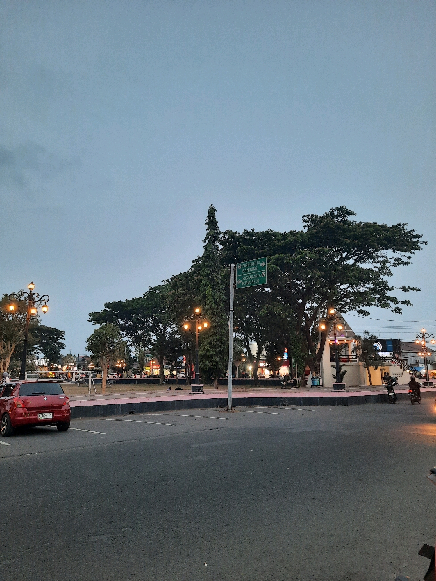 The image captures a street scene in Kutoarjo, Indonesia, likely taken during twilight hours given the overcast sky and illuminated streetlights. In the foreground, a red hatchback car is parked on the left side of the asphalt road. Beyond the road, a paved walkway is visible, bordered by a low pink-and-white wall. A large, mature tree dominates the mid-ground, its dense foliage spread wide. To the left of this tree, a tall, slender evergreen tree stands out.
A green directional signpost is positioned near the base of the large tree, indicating directions to "PURWOKERTO", "BANDUNG", "YOGYAKARTA", and "PURWOREJO". Streetlights with multiple orange lamps illuminate the area. In the background, several buildings with illuminated signs are visible, suggesting commercial activity. People are scattered throughout the park-like area, some sitting on benches, and others walking. Two motorcycles are in motion on the right side of the road, with one rider clearly visible. The overall environment appears to be a public space with trees, lighting, and pedestrian and vehicular traffic.