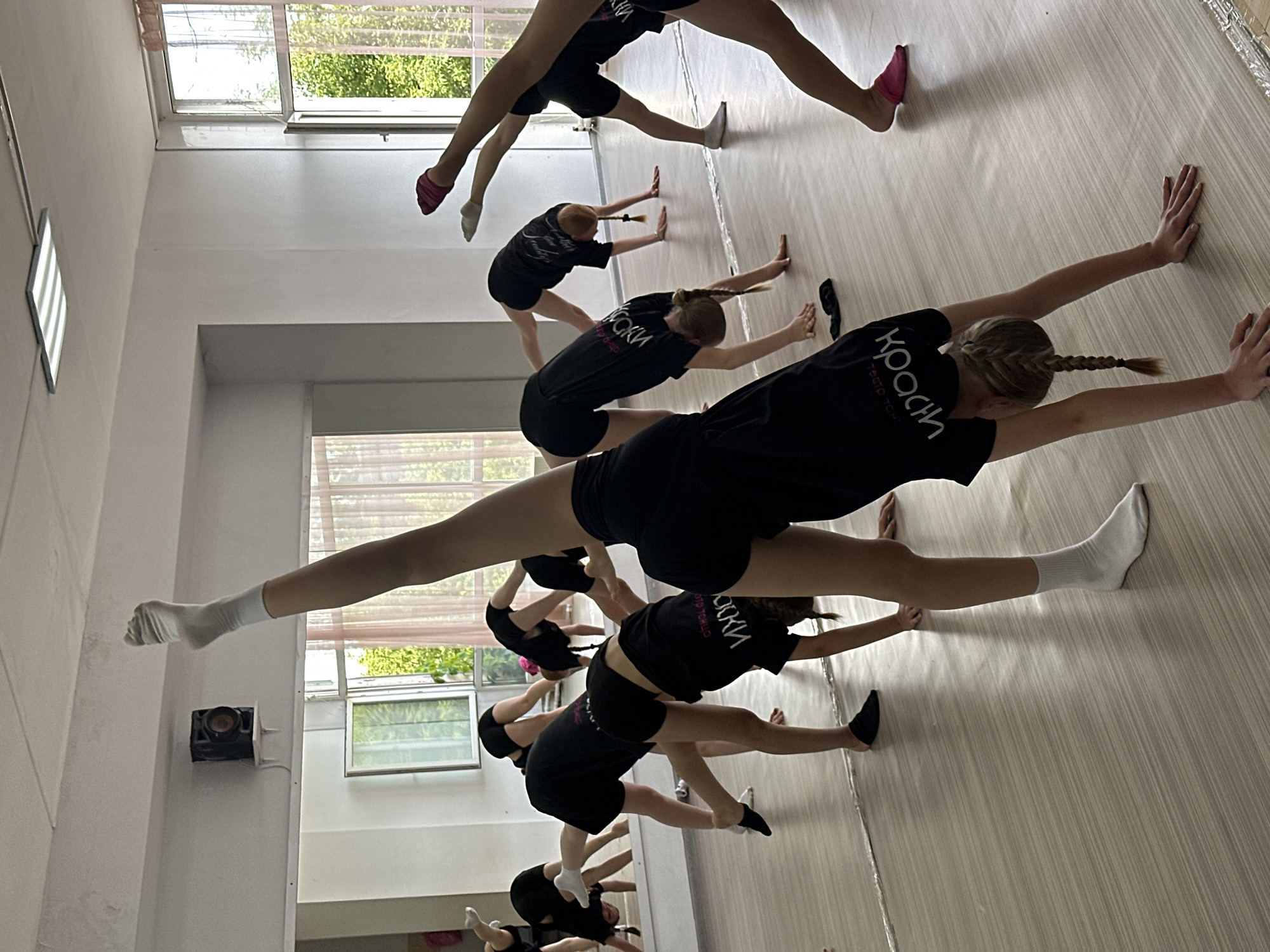 The image shows a group of young people practicing a dance or gymnastic routine in a studio. The room has a light-colored wooden floor and large windows that allow natural light to enter, suggesting it is daytime. There are several people, mostly girls, dressed in black shorts and black t-shirts with white lettering. The t-shirts have the word "КРАСИ" visible, which is Russian for "beautiful." Some of the individuals are in the process of stretching, with one person in the foreground performing a split with their legs extended wide apart. Others are seen with their hands on a wall or the floor, also stretching. The overall atmosphere appears to be one of focused physical activity and training. The location is Votkinsk, Russia.