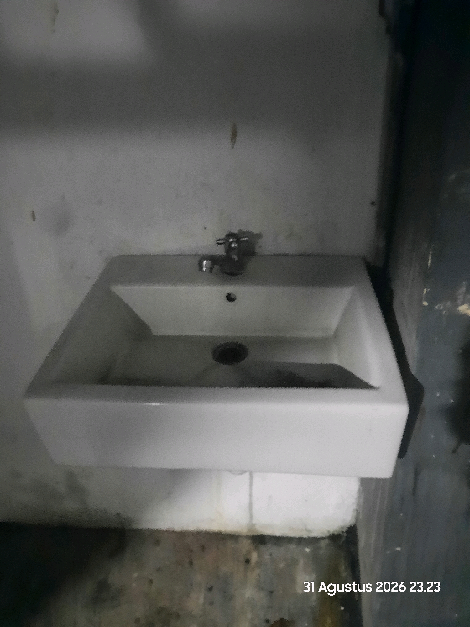 A white rectangular sink with a chrome faucet is centrally positioned against a light gray wall in what appears to be a restroom. The sink is empty, and a dark circular drain is visible at its bottom. To the right of the sink, a dark blue wall is partially visible, and the floor in front of the sink is a mottled gray and brown. In the lower right corner of the image, text reads "31 Agustus 2026 23.23". The scene is dimly lit, suggesting an indoor environment possibly in Gending, Indonesia, given the date format. There are no people or ongoing actions depicted.