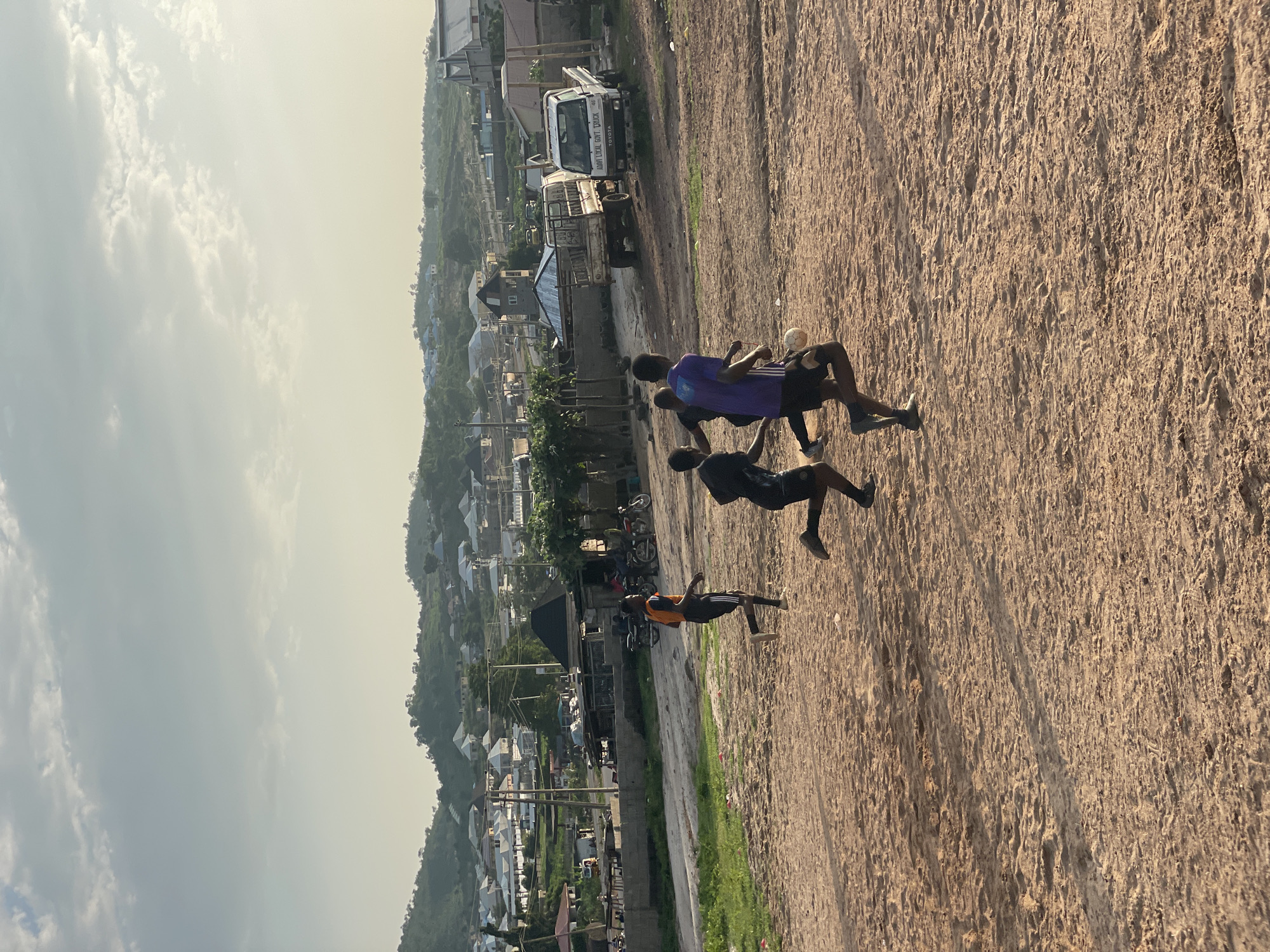 Four young men are playing soccer on a dirt field in a residential area of Lokoja, Nigeria. The field is in the foreground, with a scattering of small rocks and footprints in the reddish-brown dirt. In the middle ground, there is a village with houses and shops, some with corrugated metal roofs. In the background, hills covered with trees and more houses rise against a hazy sky. The sky is filled with soft, white clouds.
The four men are in motion, engaged in a game of soccer. One man, wearing an orange shirt and black shorts, is running towards the right side of the frame. Two other men are closer together, one in a purple shirt and black shorts, and the other in a black shirt and black shorts, both appearing to be actively playing the ball. A fourth man, also in black shorts, is partially visible running towards the left. A white soccer ball is in play between the men.
In the background, several vehicles are visible, including a white Toyota truck with lettering on its side, a flatbed truck, and several motorcycles parked near some buildings. The overall scene depicts a casual outdoor activity in a developing urban environment. The lighting suggests it is either early morning or late afternoon, with the sun casting long shadows across the field.