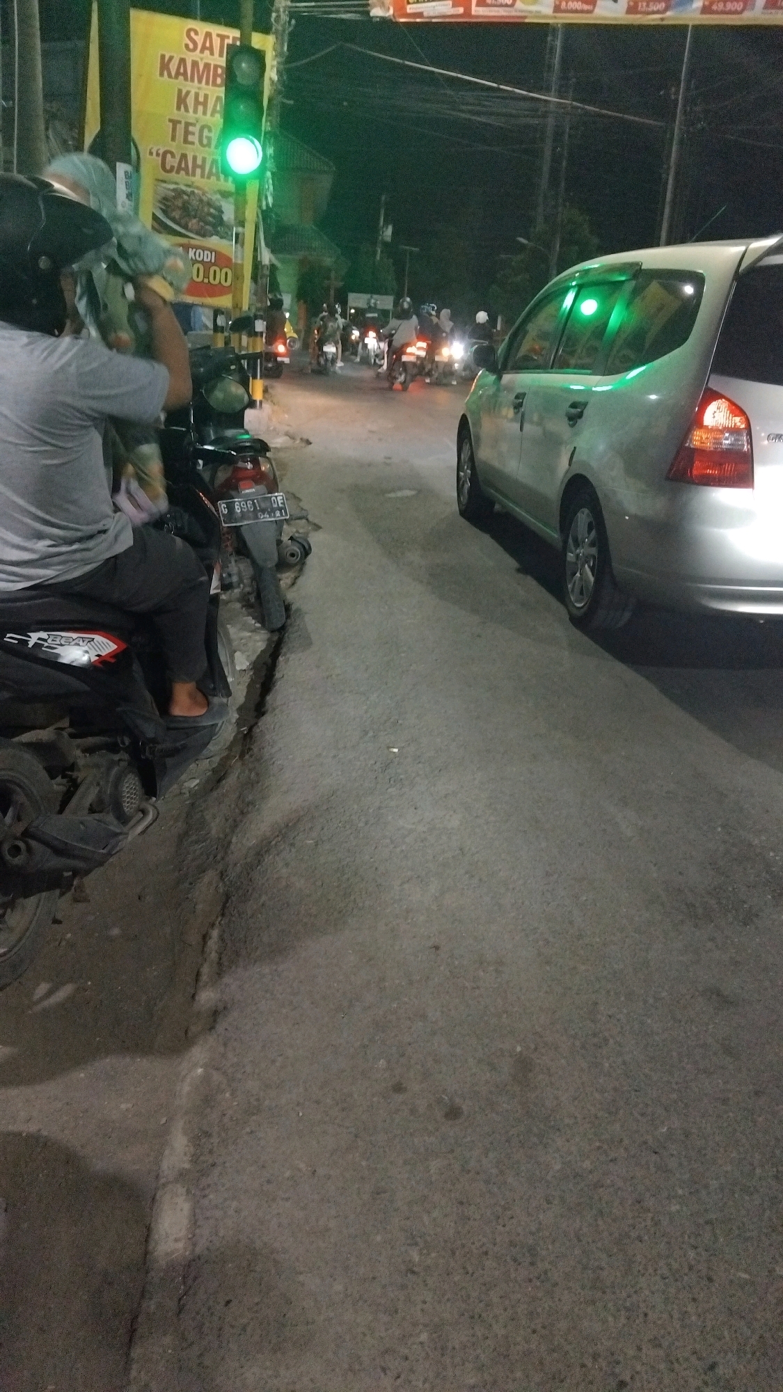 A traffic intersection in Tegal, Indonesia, is captured at night. A traffic light displays a green signal. In the foreground, a person is seated on a motorcycle, facing away from the camera. The motorcycle is parked on the side of the road, next to a cracked concrete curb. Behind the motorcycle, a silver car is visible, its interior illuminated by the green traffic light. Several other motorcycles and their riders are seen in the background, waiting at the intersection. A yellow sign with Indonesian text, possibly advertising food, is mounted on a pole to the left of the traffic light. The road surface appears to be asphalt, and the environment is dimly lit by streetlights and vehicle headlights.