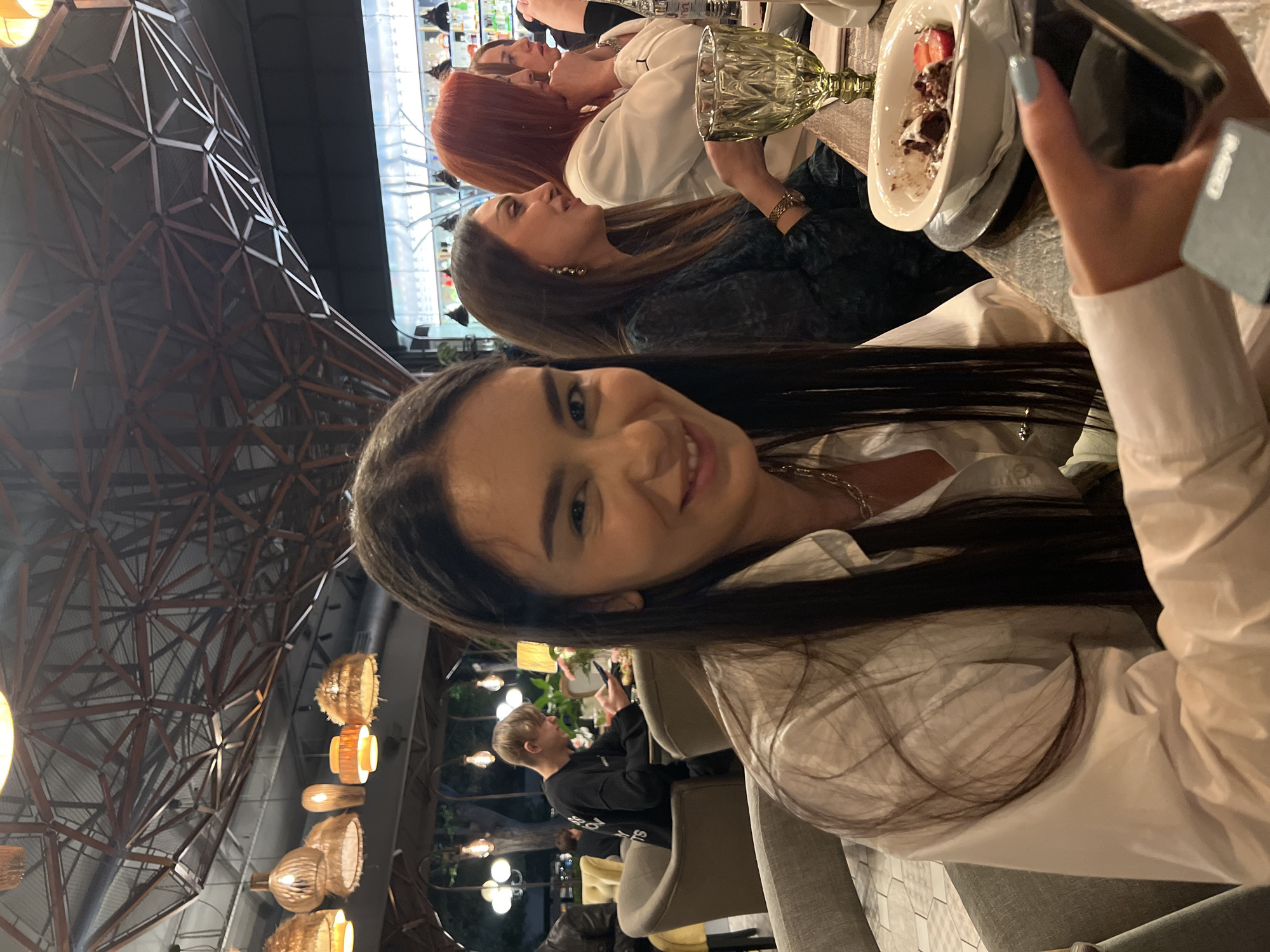 The image shows a group of four women at a table in a restaurant. The woman in the foreground is smiling and looking at the camera, holding a phone in her hand with a dessert in front of her. Another woman with brown hair and a black top is visible next to her. A woman with red hair is visible further down the table, along with another woman in white. The restaurant appears to be located in Yerevan, Armenia, based on the architectural details in the background. There are hanging decorative lights and a large metal structure.