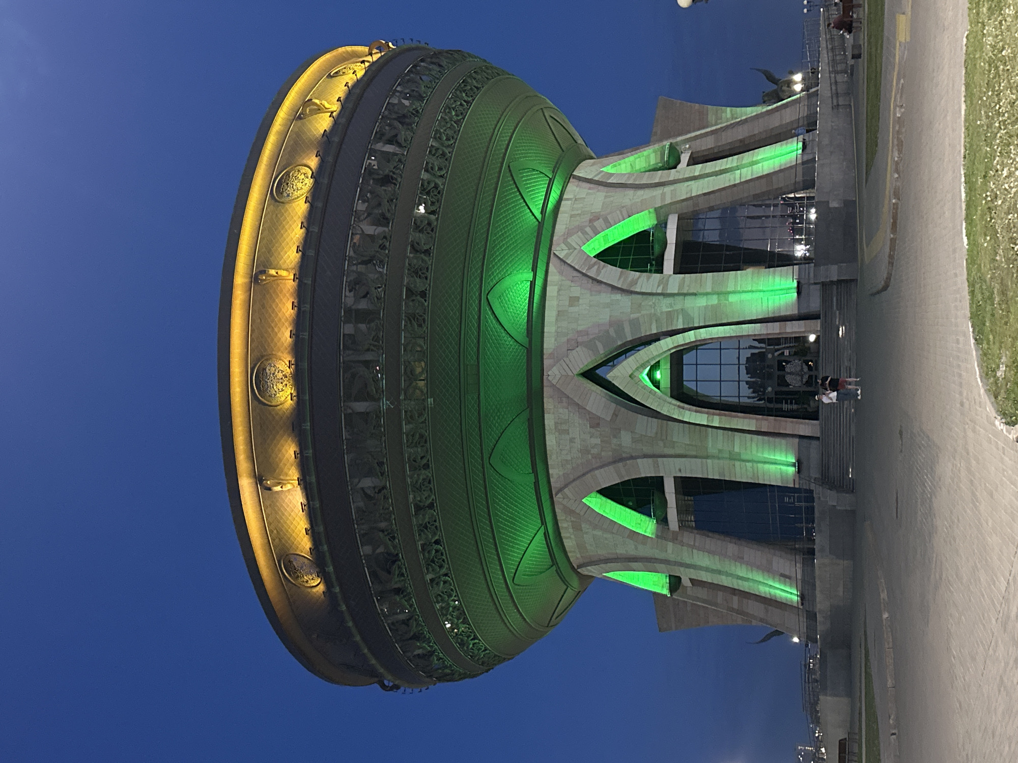 The image depicts the Kazan Family Center building, situated in the city of Kazan, Russia, at dusk. The architectural structure features a large, bowl-shaped top, illuminated in green and gold hues. Below the bowl, the building transitions to a multi-level design with glass windows, also accented with green lighting. Pavement and landscaping are visible at the base of the building, and a few people are seen near the entrance. The sky overhead is a deep blue.