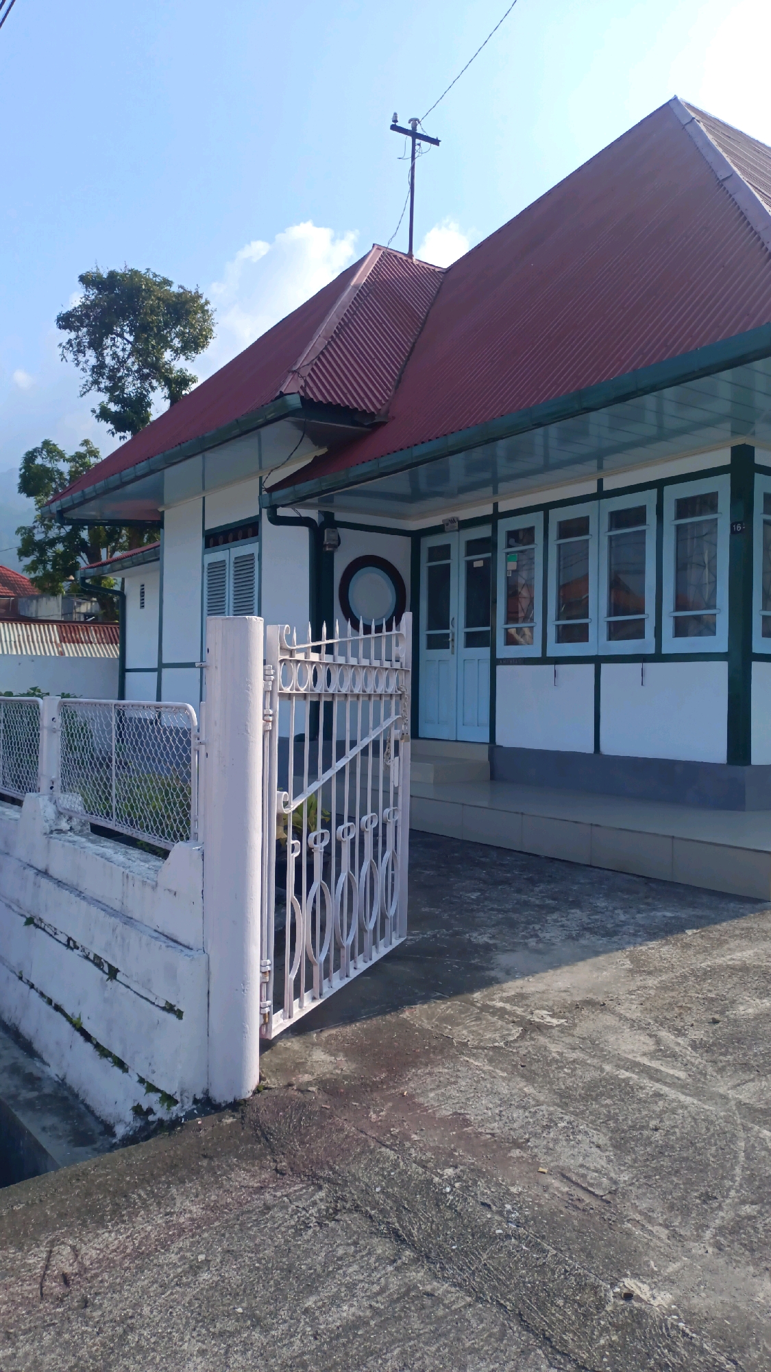 The image shows a white house with a red corrugated metal roof, a green trim, and light blue window frames and doors. A white picket fence and gate are in the foreground, with a concrete pathway leading to the house. There is a tree and a mountain in the background, under a clear blue sky with some clouds. The house is located on Jalan Jenderal Sudirman in Padang Panjang, Indonesia, a street known for its historical Dutch colonial architecture, which this house appears to be an example of.