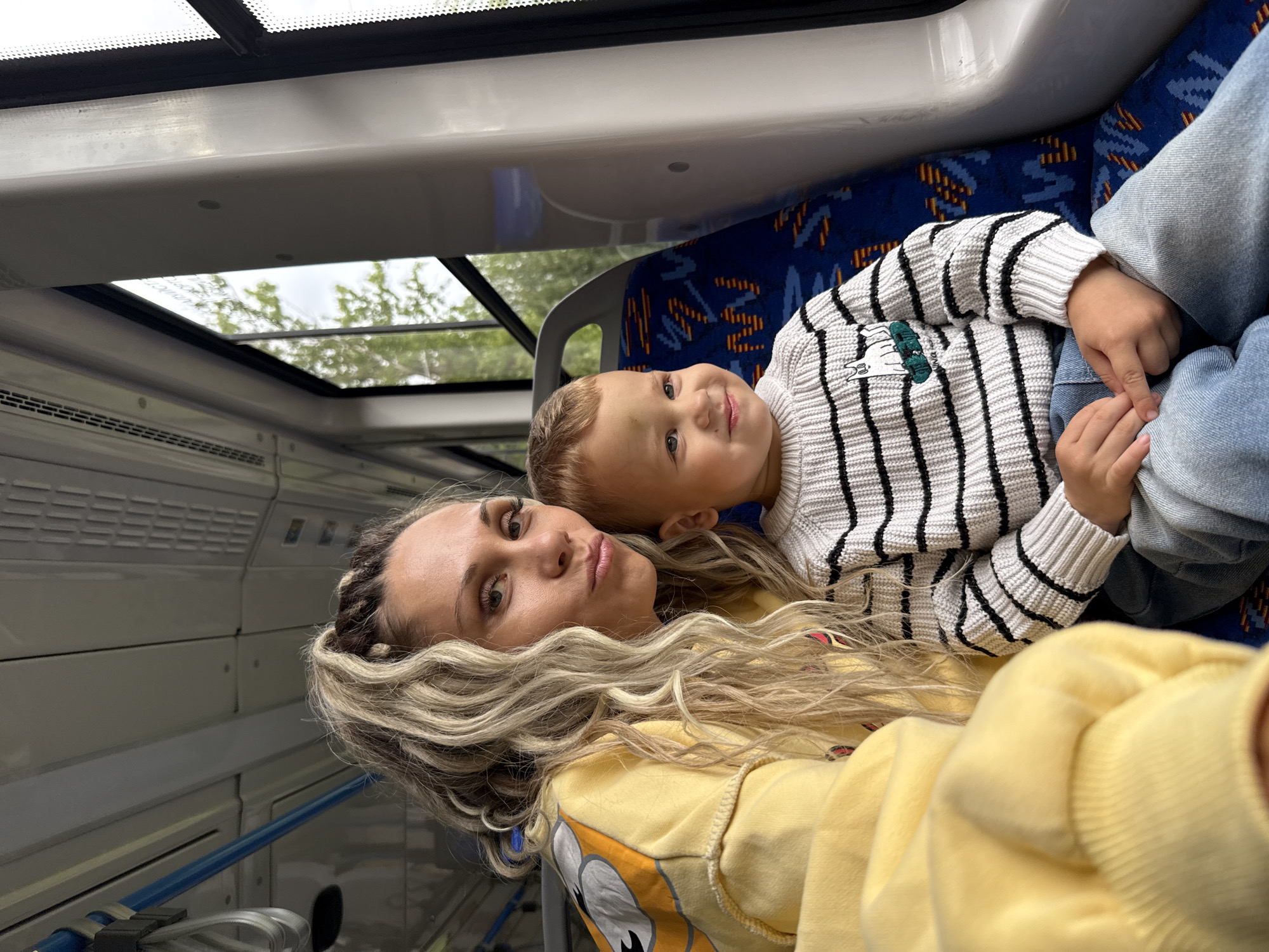 A woman and a young boy are seated inside a public transportation vehicle. The woman, with her hair styled in blonde braids, wears a yellow sweatshirt with a cartoon character design. She is looking at the camera with her lips pursed. The boy, seated next to her, is wearing a striped sweater and blue jeans. He is looking to his right with a slight smile. The seats are upholstered in a blue fabric with an orange and black geometric pattern. The interior of the vehicle appears to be a bus or a tram, with windows to the right showing a blurred view of green foliage. The overall scene suggests a casual journey. The location is identified as Rusanovo, Russia.