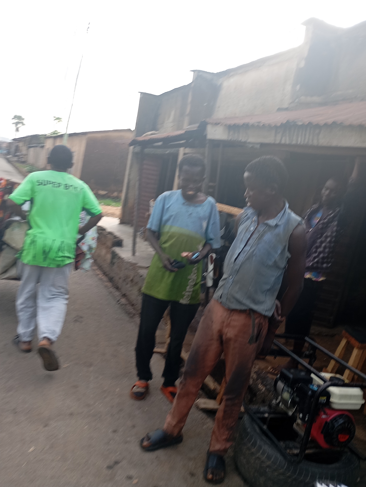 The image depicts a street scene in Shendam, Nigeria, with several individuals present. In the foreground, a person wearing a bright green t-shirt with the text "SUPER BIKES" is walking away from the camera. They are wearing light-colored pants and dark shoes. To their right, another person in a blue and green t-shirt is standing, holding a small dark object. Further to the right, a person in a light blue sleeveless shirt and orange pants is looking to their left. Behind them, another person is partially visible.
The setting appears to be an outdoor area with buildings in the background that seem to be shops or residences, constructed with corrugated metal roofs and rough walls. The ground is a paved surface, likely a road or pathway. There is some debris and equipment, including a tire and what looks like a small engine, to the right of the frame, suggesting a work or informal market area.
The lighting suggests it might be daytime, possibly overcast or late afternoon, given the overall brightness and slightly muted colors. There are no strong shadows. The activity appears to be casual, with people standing and walking around. The expressions are not clearly discernible due to the image quality and distance. The overall impression is a candid snapshot of everyday life in a Nigerian town.