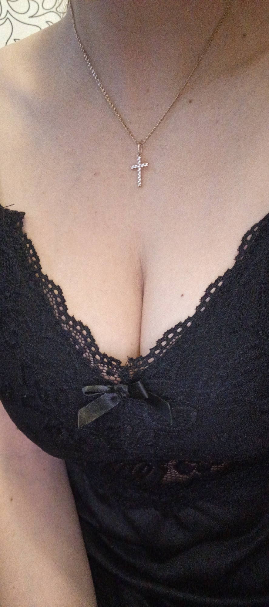 A close-up, high-angle shot captures the upper chest and décolletage of a person, located in Chaikovsky, Russia. The individual is wearing a black garment, likely a camisole or top, adorned with intricate black lace along the neckline. A small, black satin bow is tied at the center of the garment, resting in the visible cleavage. Around the neck, a thin, gold-toned chain necklace is worn, featuring a small, sparkly cross pendant as its centerpiece. The skin tone appears fair, with a few small moles visible on the chest and neck. The background is softly blurred, showing a light-colored, patterned surface in the upper left corner, indicating an indoor setting. No specific activity or emotion is discernible beyond posing for the photograph. No text is visible in the media.