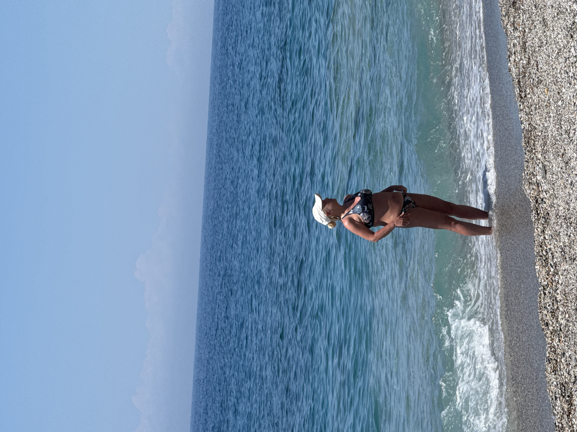 The image shows a beach scene on a sunny day in an unknown location. A woman wearing a swimsuit and a hat is standing in the water at the edge of the beach. The water appears clear and calm. The beach is made of small pebbles. The sky is blue with some clouds visible in the distance. The image is taken from a high angle and is oriented vertically.