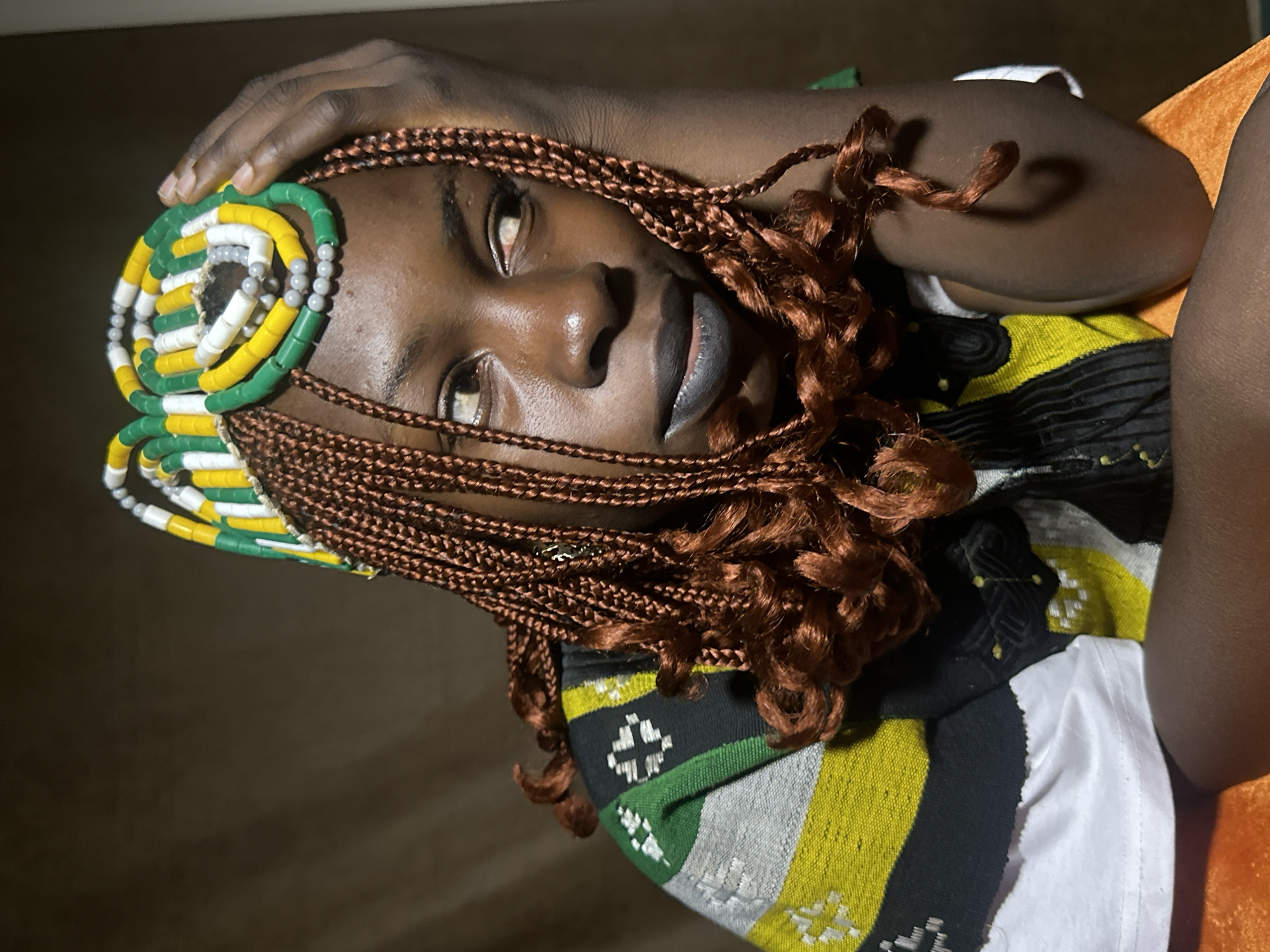 The image shows a close-up portrait of a young Black woman with dark skin and braided hair. She is wearing a colorful, beaded headdress and a patterned garment with green, yellow, black, and white elements. Her right hand is resting on her head, and she is looking away from the camera. The background is a muted brown, suggesting an indoor setting. The lighting creates a dramatic effect, with highlights on her skin and a shadow cast behind her. The overall impression is one of artistic expression and cultural adornment. There is no information in the image to suggest it was taken in Jos, Nigeria, or any other specific city.