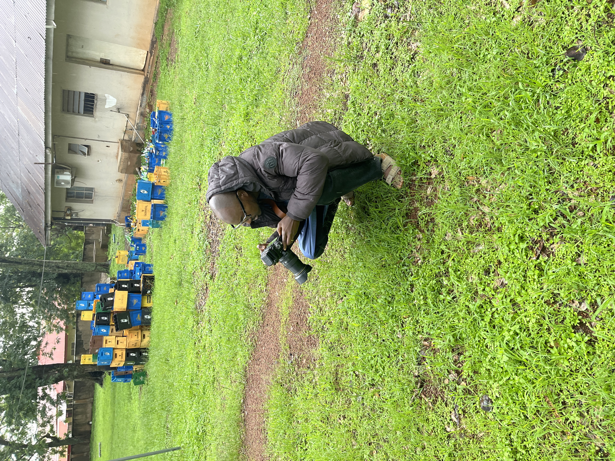 A bald Black man wearing glasses and a hooded puffer jacket crouches in a grassy outdoor area, holding a camera with a lens attached to his right hand. He is looking at the camera as if to take a picture. The ground is covered in green grass, with a brown dirt path visible to the left. In the background, there is a large pile of blue, yellow, and black plastic crates, some stacked high. Beyond the crates, a building with a corrugated metal roof and a light-colored facade is visible. The overall scene suggests an outdoor location, possibly a compound or grounds of a facility, in Jos, Nigeria, where the man is engaged in photography amidst a collection of beverage crates.
