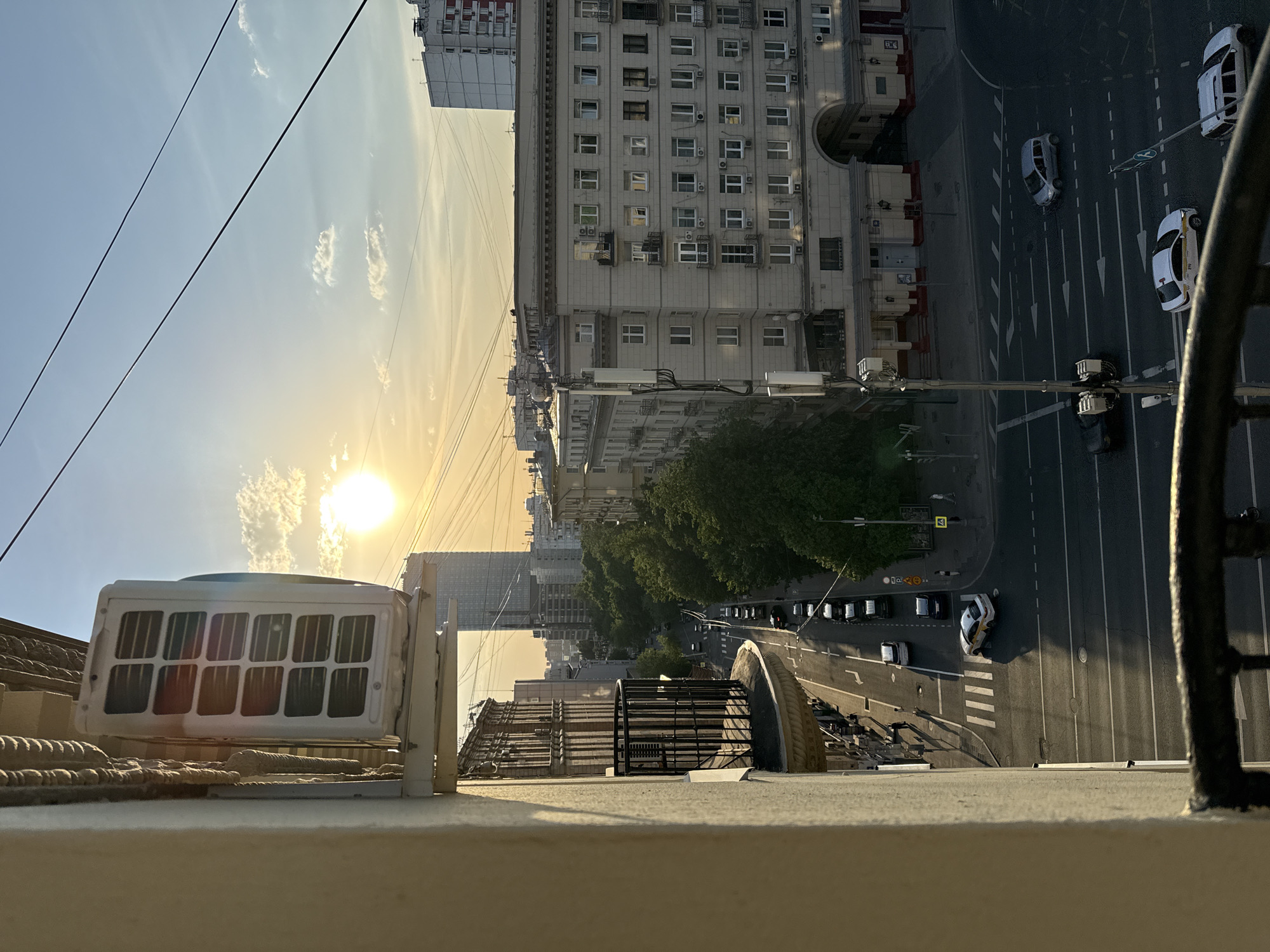 From a high vantage point in Moscow, Russia, the sun is setting over a cityscape. The street below shows vehicles in transit and parked. Close to the viewer are architectural elements like railings and mechanical equipment. Dominating the view are buildings of varying heights, with a row of trees adding a touch of greenery to the urban scene.