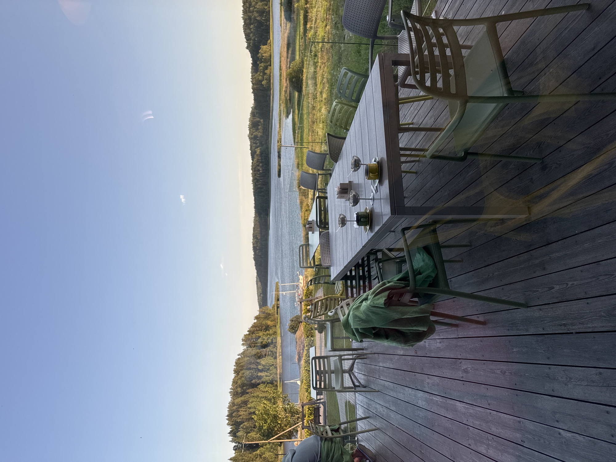 The image depicts an outdoor seating area at a restaurant with a view of a lake and surrounding trees. The foreground shows a wooden deck with several tables and chairs. One large table is set with two cups and saucers, and two wine glasses. Green blankets are draped over some of the chairs. In the background, the lake is calm, with several sailboats anchored. The shore is lined with dense green trees. The sky is clear and blue, indicating it is daytime. The location is stated to be the city of Kurkijoki, Russia.