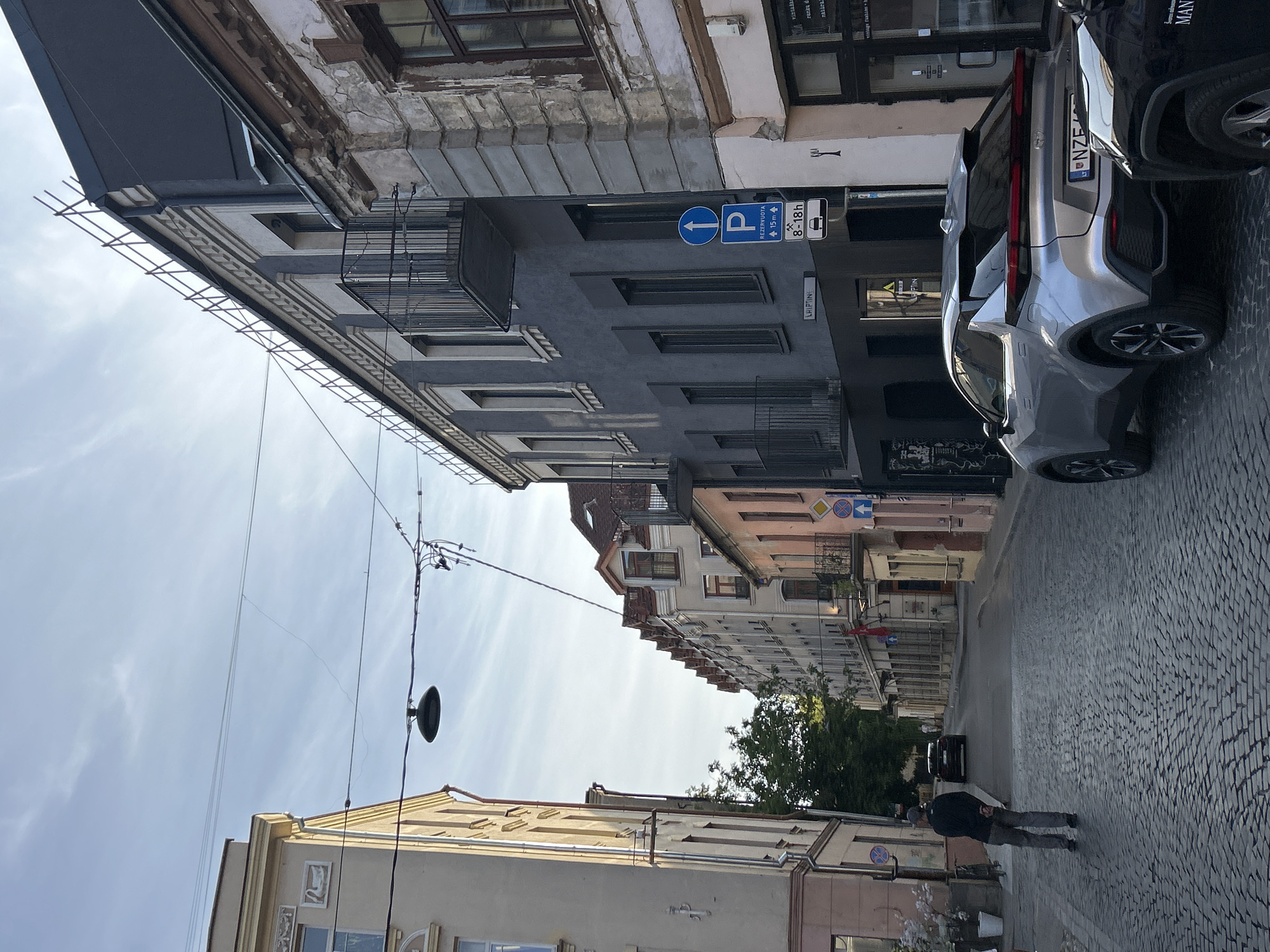 The image depicts a street scene in Vilnius, Lithuania, characterized by cobblestone paving and a row of multi-story buildings. The architecture suggests an older European city. The sky is clear with some wispy clouds, and the lighting indicates it is either early morning or late afternoon.
On the left side of the street, a pale yellow building with decorative elements on its upper facade stands prominently. Further down the street, several buildings of varying colors, including pink and gray, line the left side, some featuring balconies with wrought-iron railings. The right side of the street is dominated by a modern-looking dark gray building with a contrasting lighter gray section and balconies. An older, weathered building with visible cracks and peeling paint is adjacent to it.
In the foreground, a person wearing a dark jacket and gray pants is walking away from the camera down the cobblestone street. A silver SUV is parked on the right side of the street, facing away from the camera, with its license plate partially visible. A portion of a black vehicle is visible to the right of the silver SUV.
Street furniture includes a street lamp hanging from wires, several traffic signs, and what appear to be potted plants or decorative displays near the entrance of one of the buildings on the left. One sign, in particular, indicates parking restrictions with "REZERVUOTA" and a time frame of "8-18h". The street itself is relatively narrow, and further down, trees can be seen behind the buildings. The overall impression is a quiet urban street in a historic part of the city.