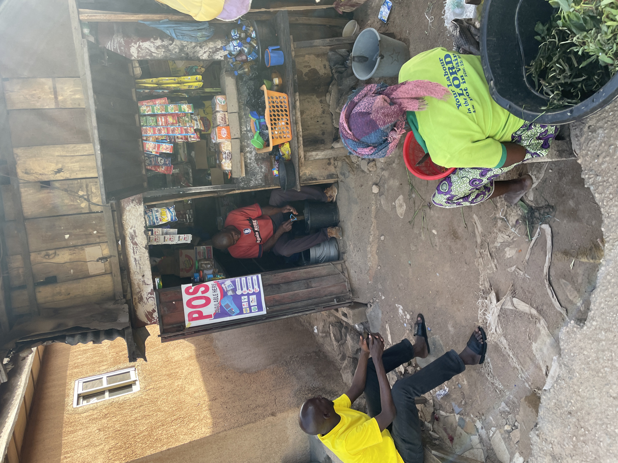 The image shows an outdoor scene in Jos, Nigeria. In the foreground, a man in a yellow shirt and black pants sits on the ground. Behind him, another man in a red shirt sits in a small shop. The shop has various items on display, including snacks and drinks. A "POS AVAILABLE HERE" sign is visible next to the shop's entrance. To the right, a woman wearing a green shirt and headscarf is sitting near a bucket filled with green plants. A second bucket and various other objects are near her. The environment appears to be a semi-urban setting with a building visible behind the shop.