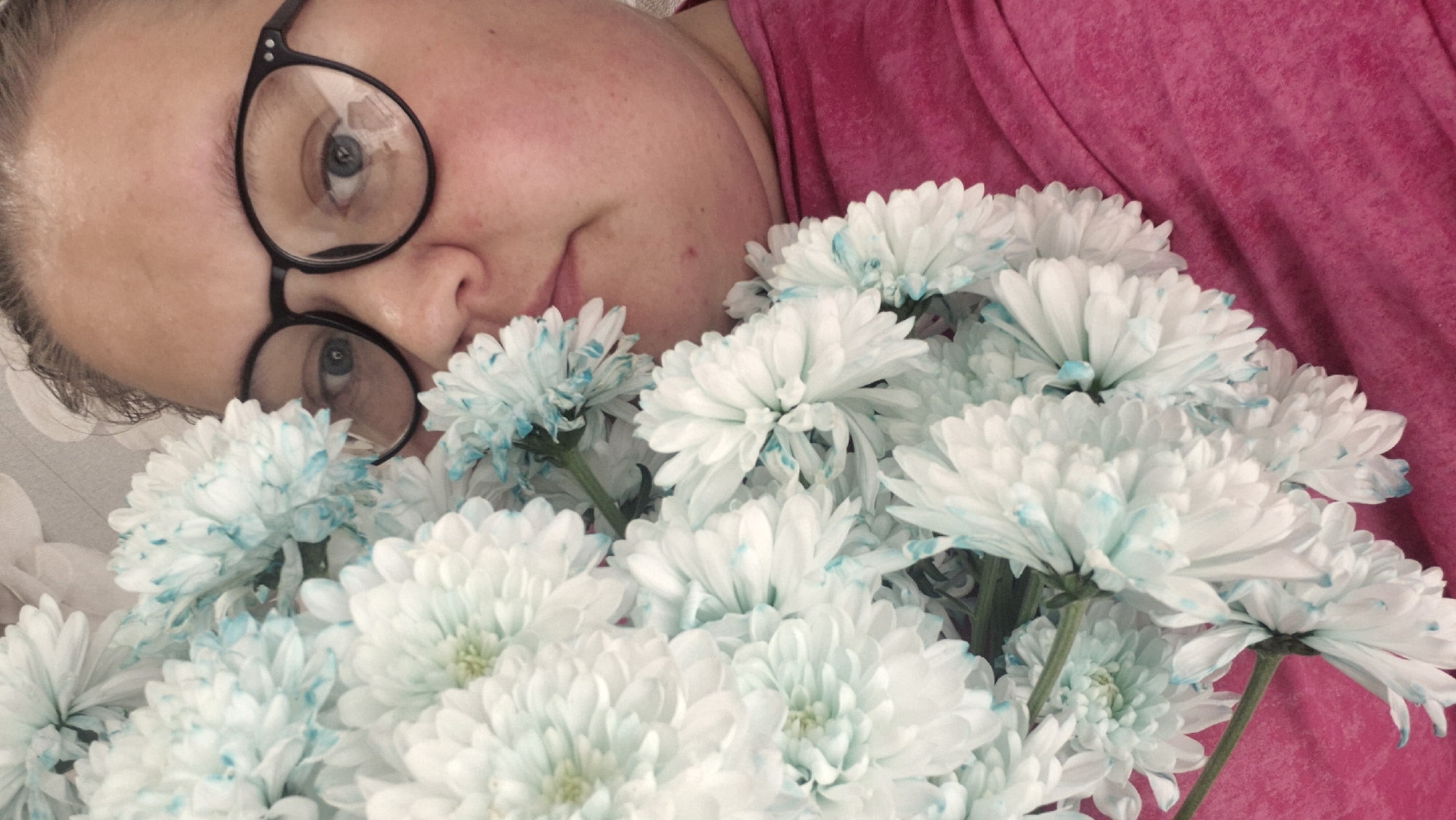 A person with fair skin and wearing black-rimmed glasses is lying down and holding a bouquet of white chrysanthemums with blue-tipped petals. The person is wearing a pink top. The setting appears to be indoors, possibly a home, based on the casual pose and the presence of fabric or bedding visible beneath the person and flowers. The image doesn't provide any information to identify the exact location in Chaikovsky, Russia.
