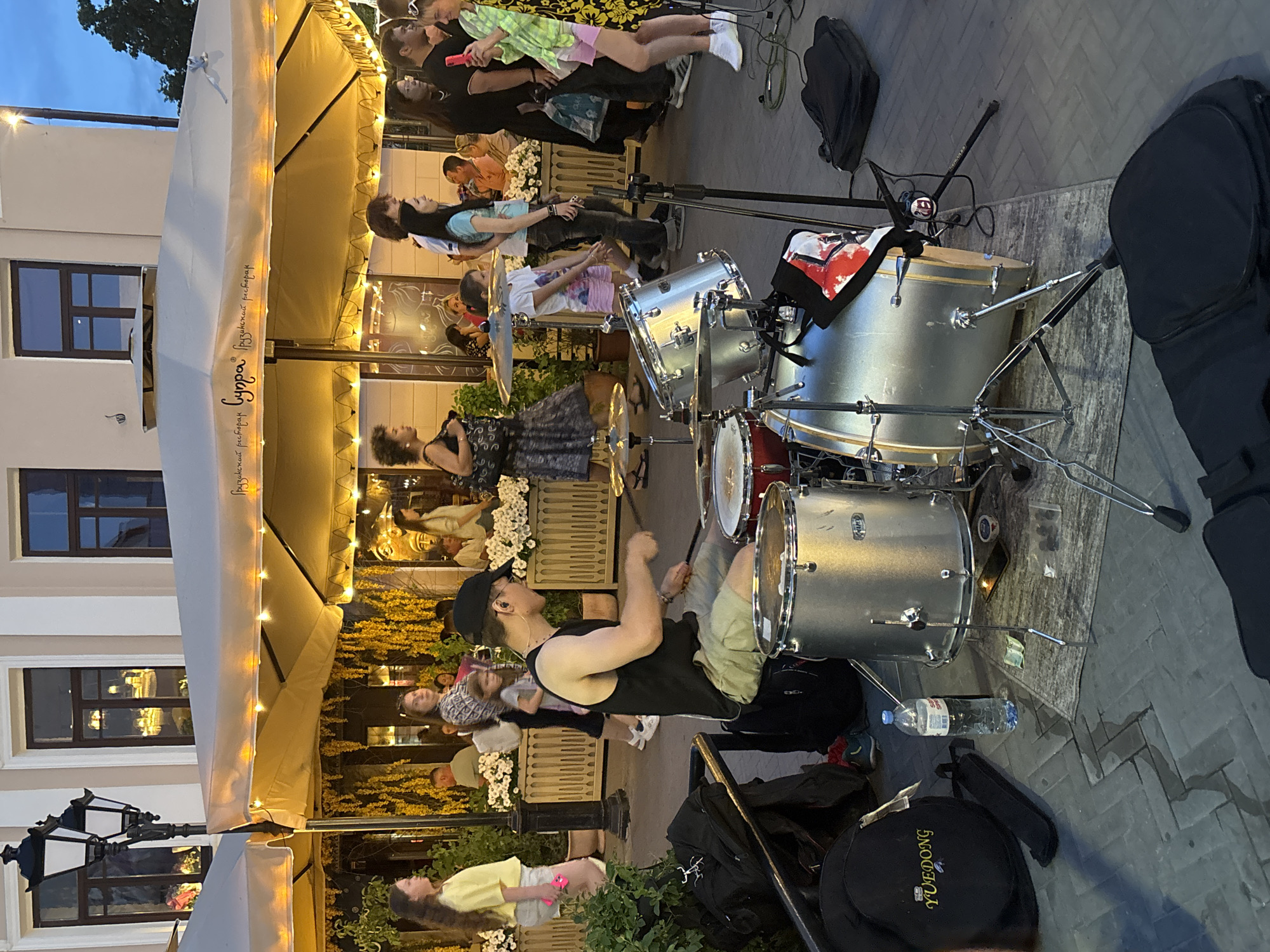This image captures a street scene in Kazan, Russia, likely in the summer evening. In the foreground, a male drummer is actively playing a drum set on a cobblestone street. He wears a black tank top, light-colored shorts, and a black baseball cap. Various parts of the drum set, including cymbals and snare drums, are visible. To the left of the drummer, a black drum bag with the "YUEDONG" logo is placed on the ground. A bottle of water and some loose change are also on the ground near the drum set.
Behind the drummer, a group of people are walking past. Some are dining under a large, cream-colored awning adorned with yellow lights, which appears to be part of an outdoor cafe or restaurant. The building behind the awning has large windows reflecting the evening sky. The overall atmosphere suggests a lively urban environment where street performers and patrons of cafes coexist.