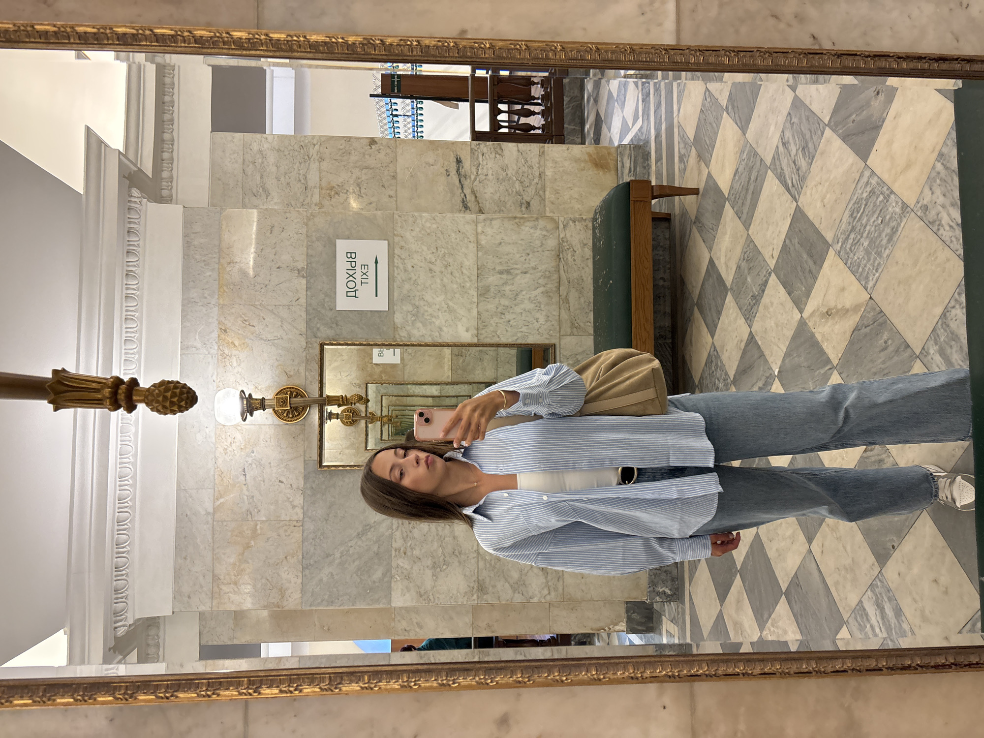 The image is a selfie taken in a mirror, depicting a young woman standing in a hallway with marble walls and a checkered floor. The woman is wearing a blue and white striped oversized shirt over a white tank top and light blue jeans. She is holding a pink smartphone in her right hand, taking the picture. A tan shoulder bag is slung over her left shoulder. She has long brown hair and is looking at her reflection.
The setting appears to be indoors, likely in a public building or an older establishment, given the ornate gold-framed mirror and the classical architectural details of the hallway, such as the decorative moldings and the marble tiling. A green upholstered bench is visible in the background. On the wall, there is a sign with an arrow pointing to the right, with the text "EXIT" and "ВРИХОД" written in English and Russian respectively. The lighting suggests it is daytime, with natural light possibly coming from an unseen source. The overall mood is casual and self-aware, typical of a mirror selfie. The floor is laid with large marble tiles in a diamond pattern of light and dark grey. The reflection in the mirror also shows other architectural elements, including what appears to be a staircase and a chandelier or ornate light fixture.