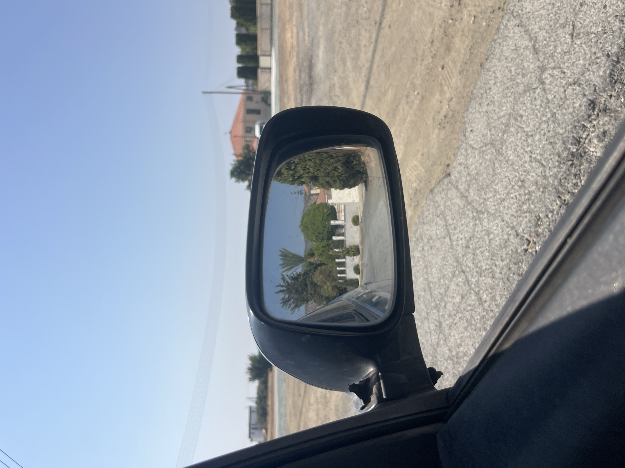 The image shows a view from inside a car, looking out the side mirror. The side mirror reflects a street scene in Parekklisia Community, Cyprus, with several houses and trees visible. The car appears to be parked on a dirt road. The sky is clear and blue.