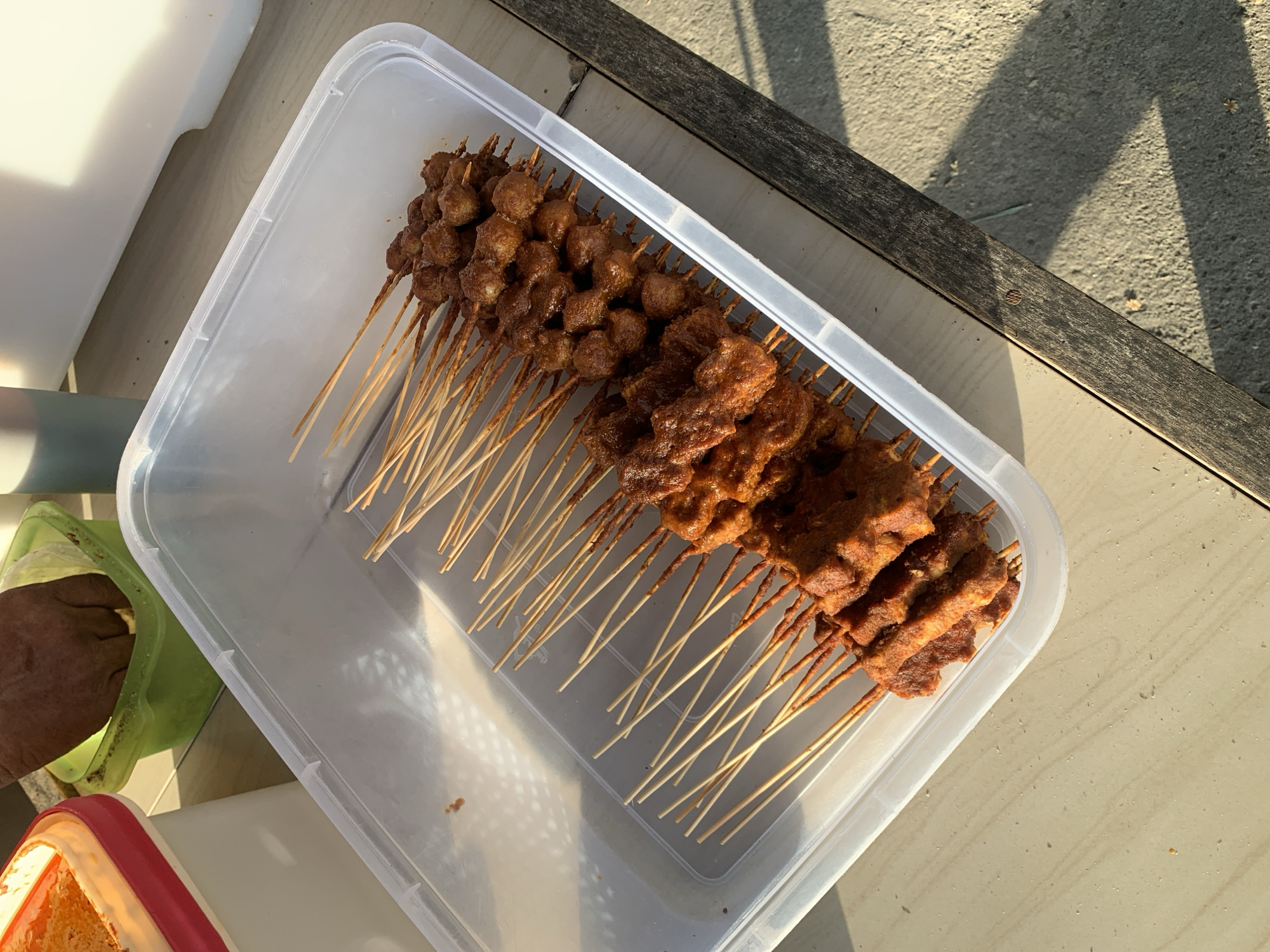 In Lhokseumawe, Indonesia, a clear plastic container filled with skewered food is placed on a wooden table. The skewers contain two types of food: a row of darker, round meatballs on the right, and a row of flatter, rectangular pieces of marinated meat on the left. The food appears to be seasoned with a reddish-brown spice mix. In the background, to the left, are two other containers. One is a white container with a red lid, partially filled with a bright orange substance. Next to it is a green container where a hand, possibly preparing the food, is dipping something into a light yellow material. The scene is illuminated by natural light, casting shadows on the table and surrounding concrete surface.
