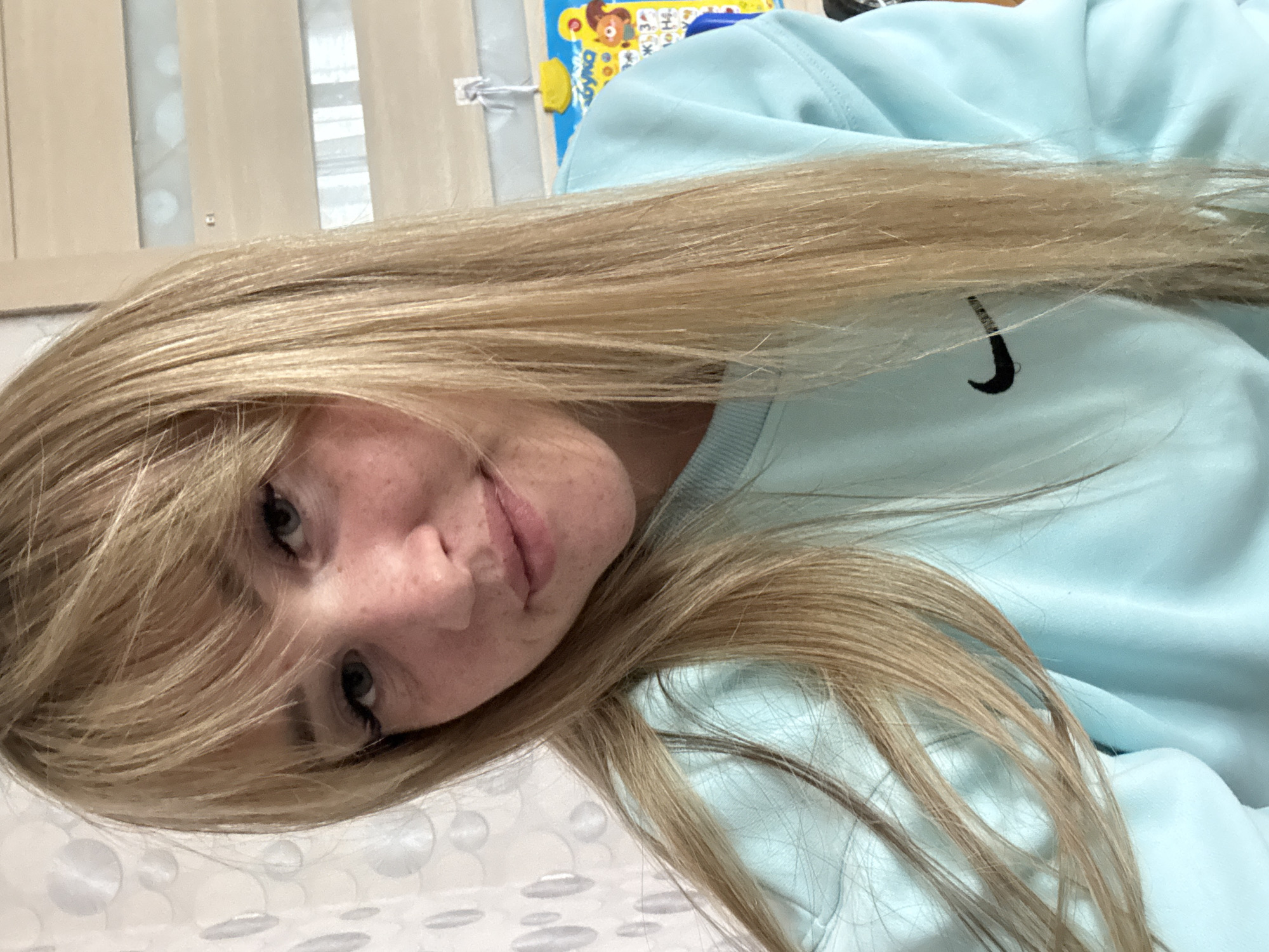 A young woman with blonde hair and freckles, wearing a light blue Nike sweatshirt, poses for a selfie indoors. Her hair is long and straight, falling over her shoulders. Her eyes are light blue, and she has on noticeable eyelashes. She is looking directly at the camera with a slight smile. Behind her, a wall with a patterned wallpaper and a wooden door or cabinet can be seen. A bright yellow and blue educational poster is also visible on the wall. The scene appears to be in a residential setting, possibly a bedroom or living area. No specific landmarks of Марковский, Russia are identifiable.