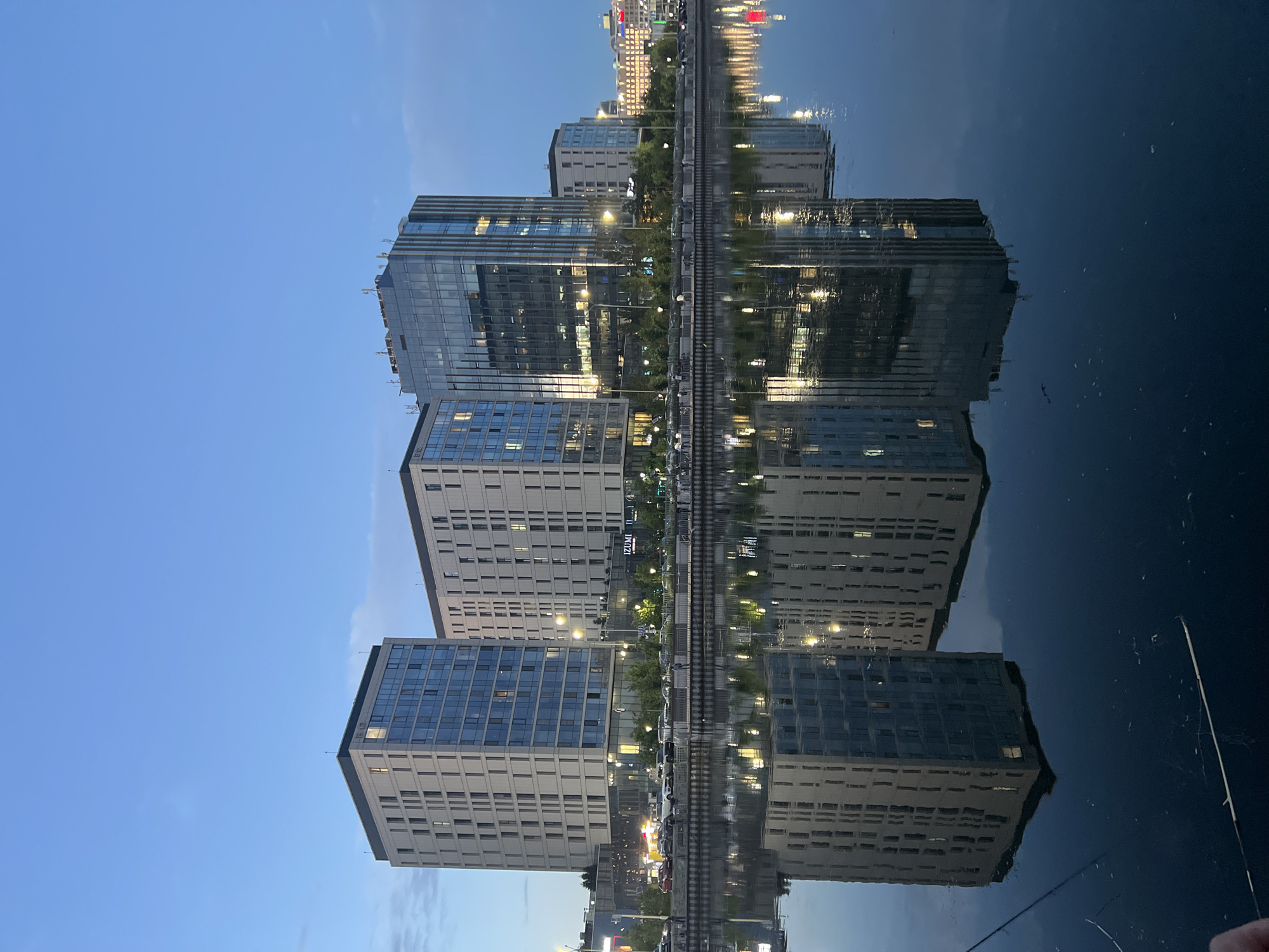 A body of water reflects modern buildings at dusk in Astana, Kazakhstan. The water surface is calm, with clear reflections of the structures on its surface. The buildings are tall, with glass facades and visible lights from within. Some buildings appear to be residential apartments, while others might be office buildings. There is a street or walkway along the waterfront, with cars parked and a few people visible. The sky is a deep blue, indicating the transition from day to night.